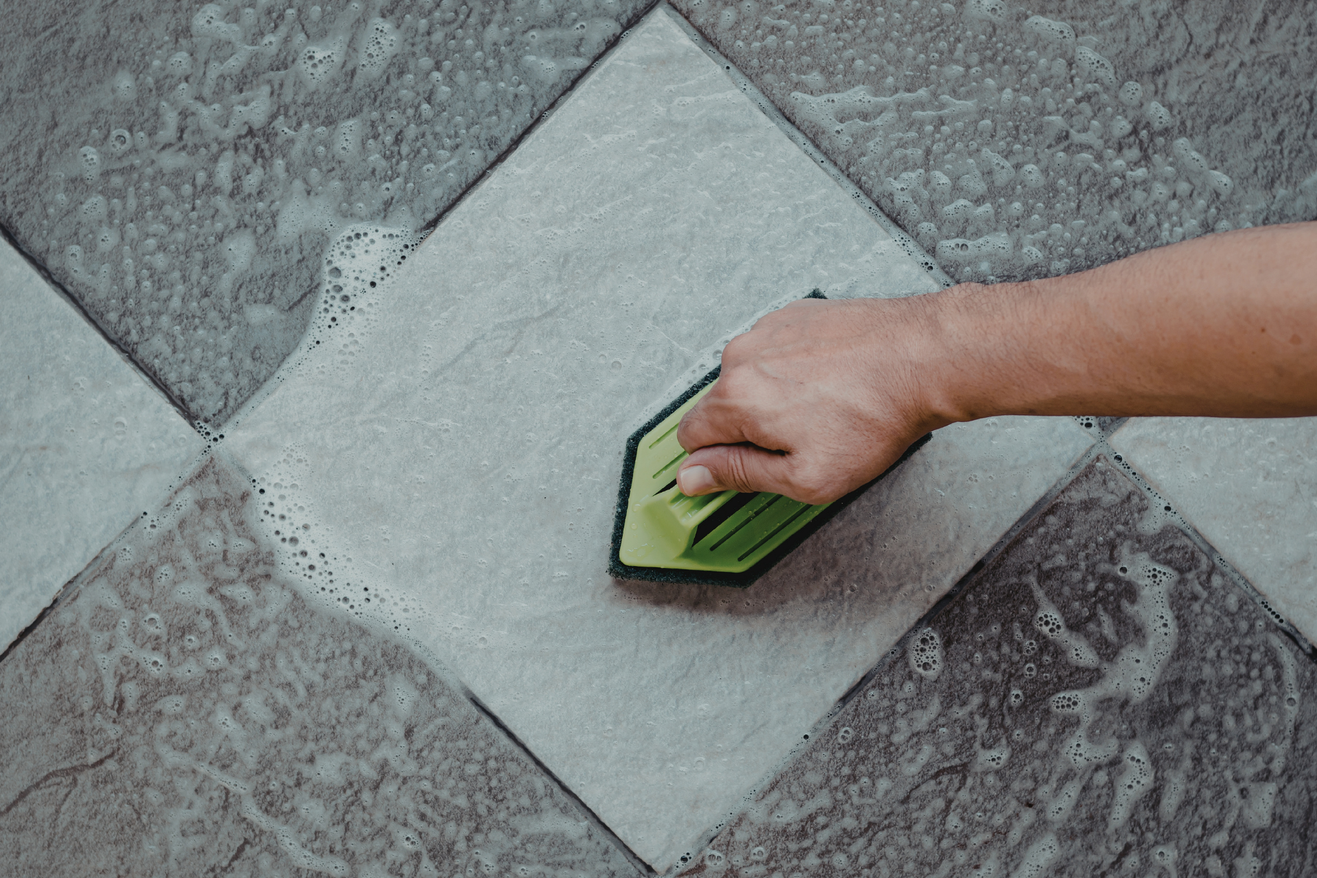 Hand using a green scraper to clean a wet tiled floor