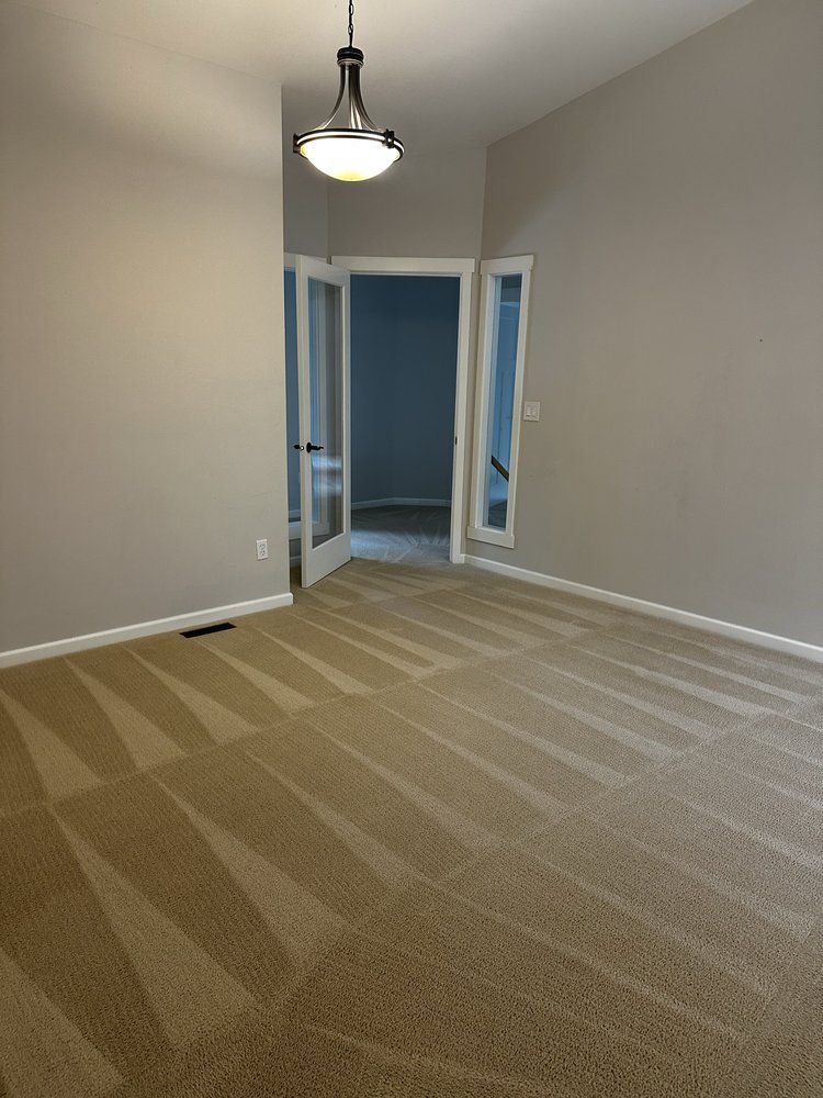Empty carpeted room with gray walls, white trim, and a hanging ceiling light, viewed toward an open doorway.