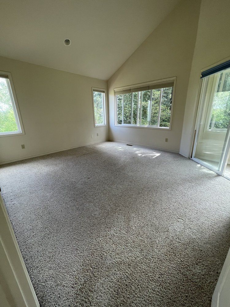 Empty carpeted living room with large windows and sliding glass door, beige walls and vaulted ceiling
