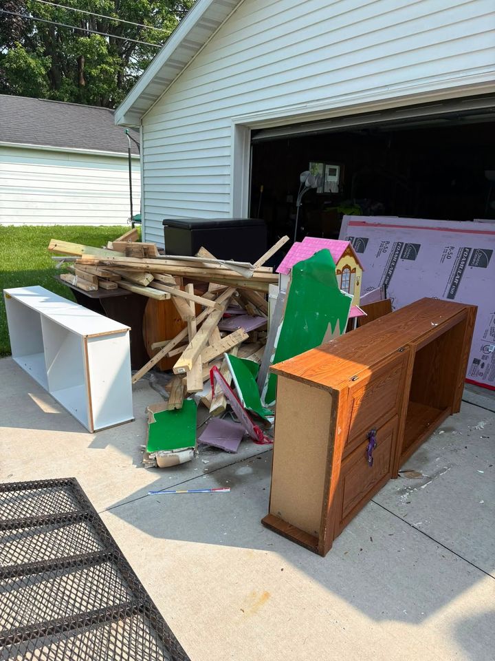 Pile of broken furniture and debris cluttering a driveway beside an open garage.