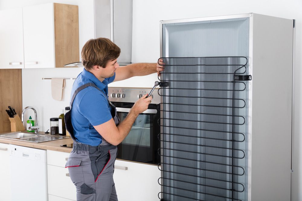 Serviceman In Overall Working On Refrigerator With Screwdriver In Kitchen — Entire Trades in Central Coast, NSW