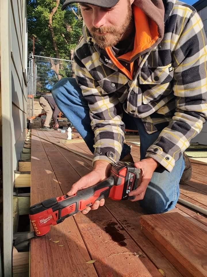 A Man Is Kneeling Down on A Wooden Deck Holding a Tool — Entire Trades in Maitland, NSW