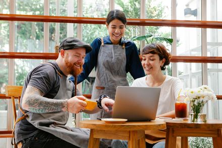 Three people sharing coffee and smiling around a laptop in a bright café