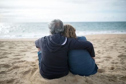 Two people sitting on a sandy beach, facing the ocean and embracing under a cloudy sky