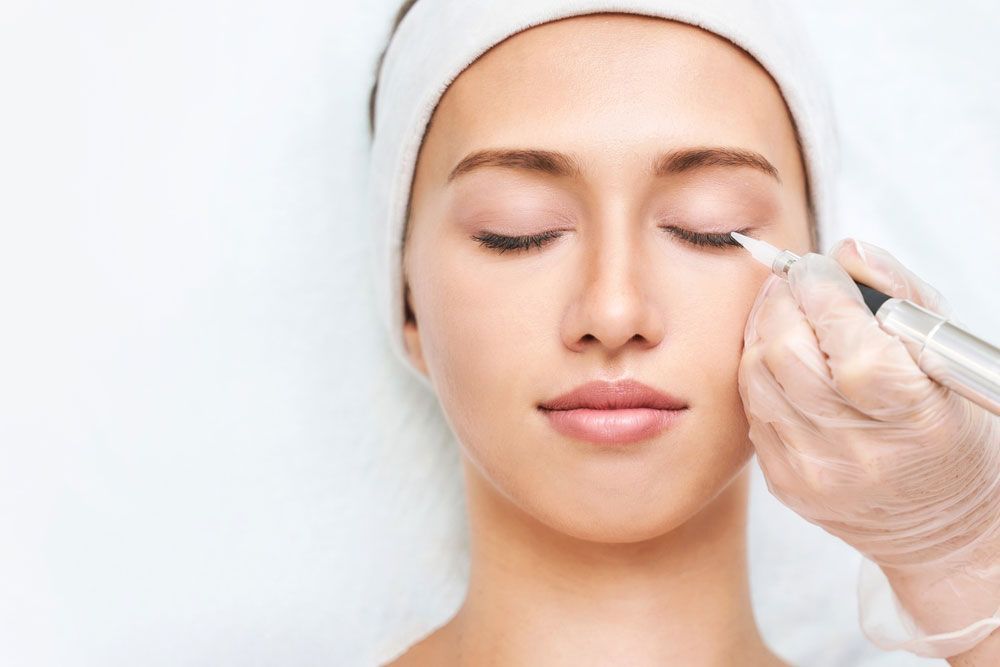 Gloved hand injecting serum under a woman’s eye as she lies with eyes closed on a treatment bed