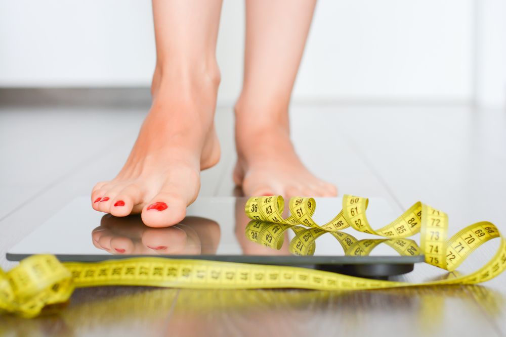 Bare feet on a scale with a yellow measuring tape, suggesting weight loss or fitness tracking
