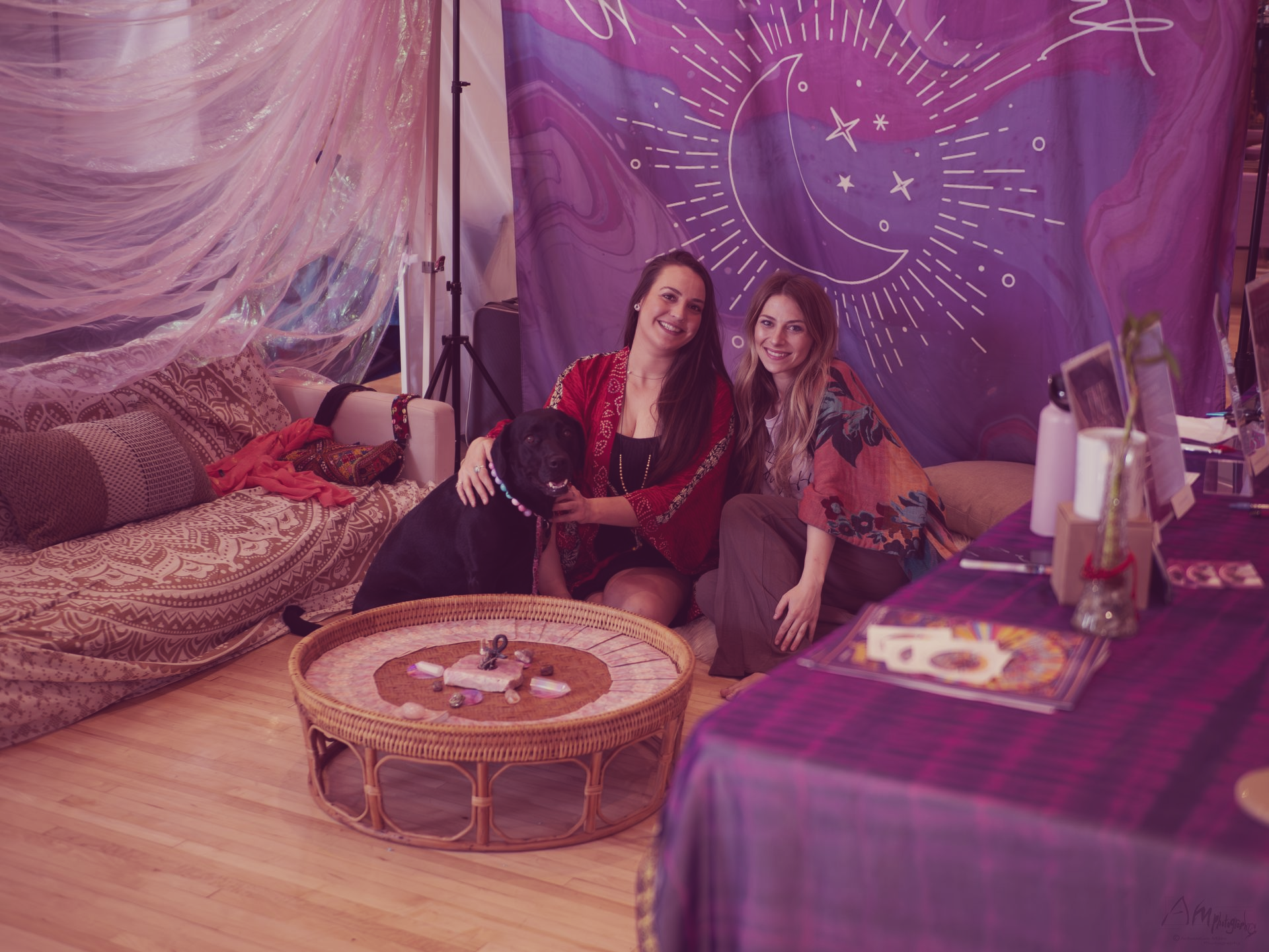 Two people sitting in a purple bohemian tent with patterned rugs, cushions, and a low table.