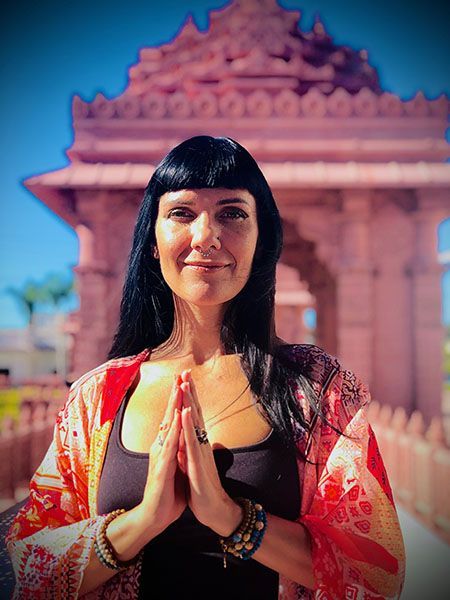 Woman with hands in prayer, in front of a temple; wearing a black top and orange shawl.