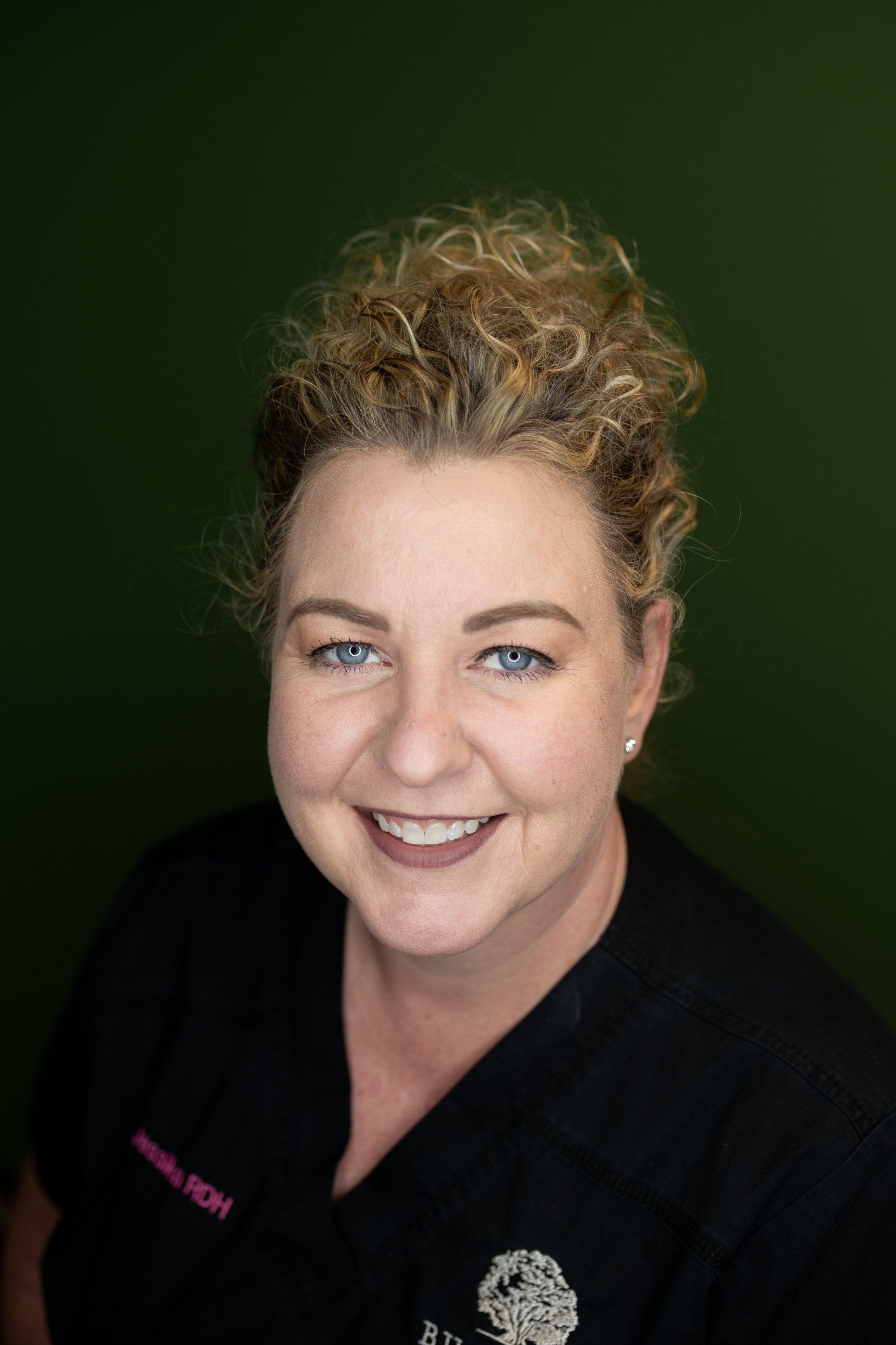 A woman with curly hair is smiling and wearing a black scrub top.