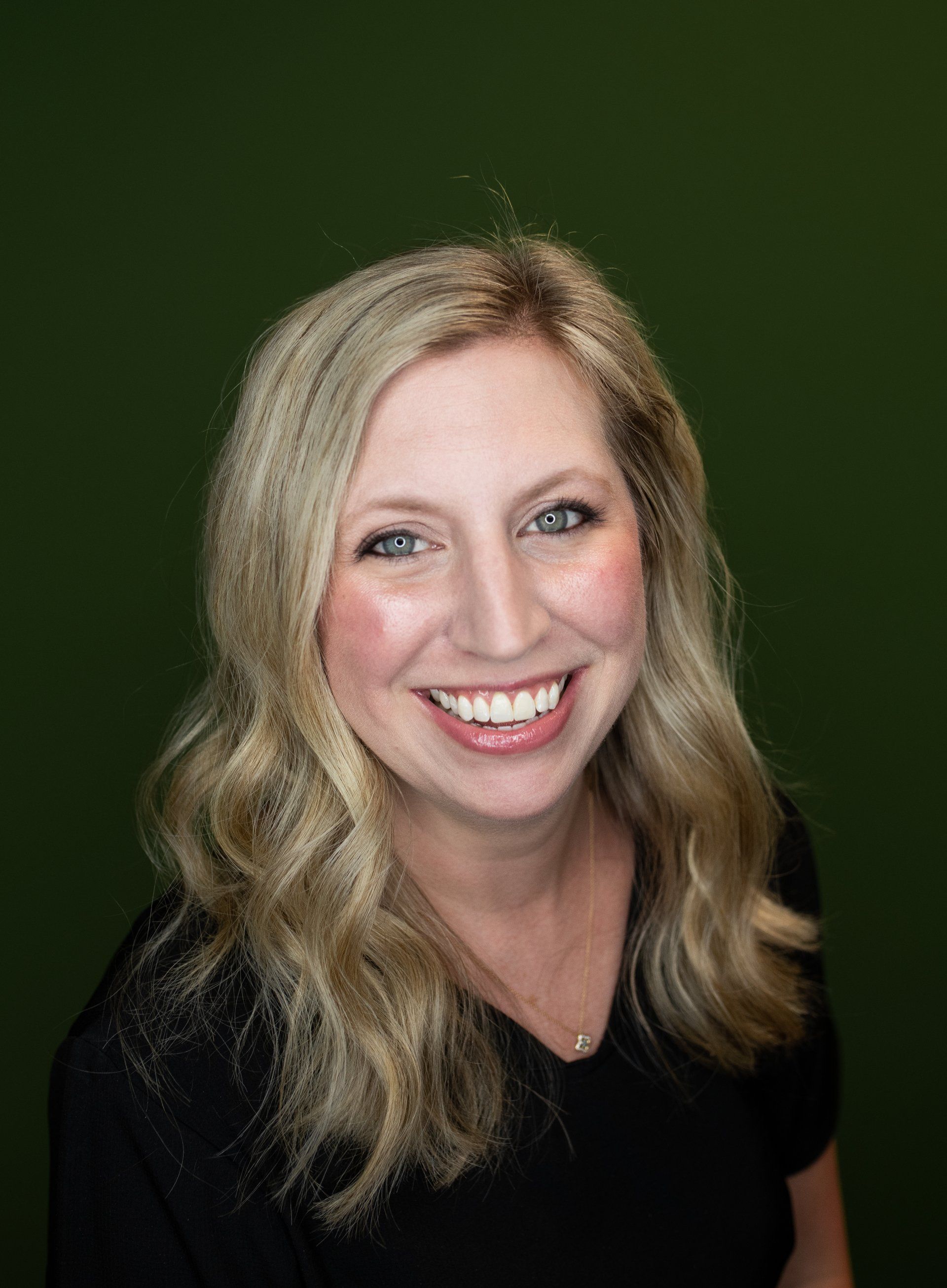 A woman in a black shirt is smiling in front of a green background.