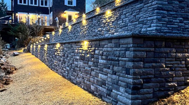 Stone retaining wall with built-in lights, gravel path, and house in the background at dusk.