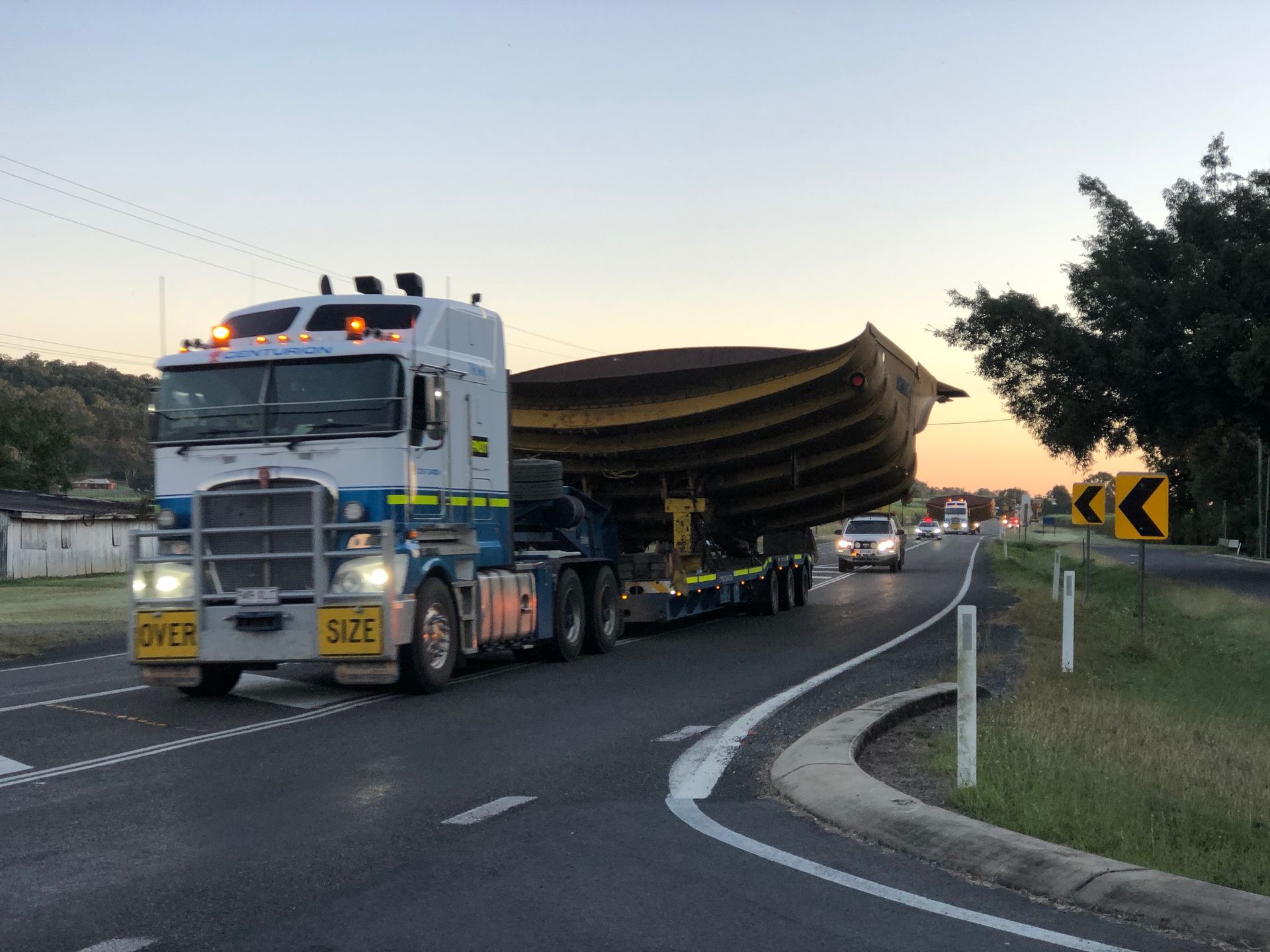 A Kenworth truck transports a large, curved metal structure on a flatbed trailer along a paved road at dusk — Col Adamson Transport Services in Farleigh, QLD
