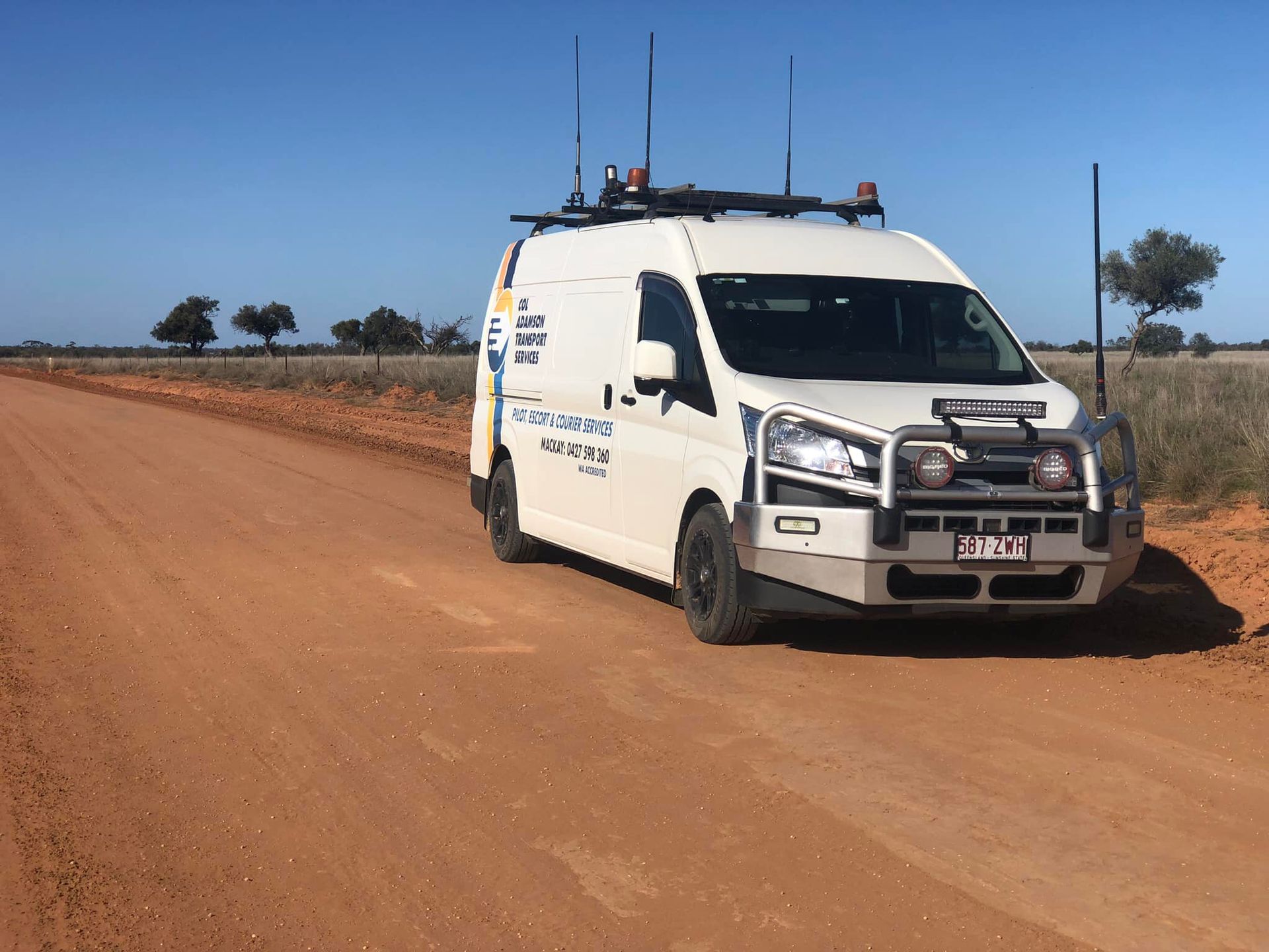 White van with antennas, driving on a red dirt road in a rural landscape under a clear blue sky — Col Adamson Transport Services in Farleigh, QLD