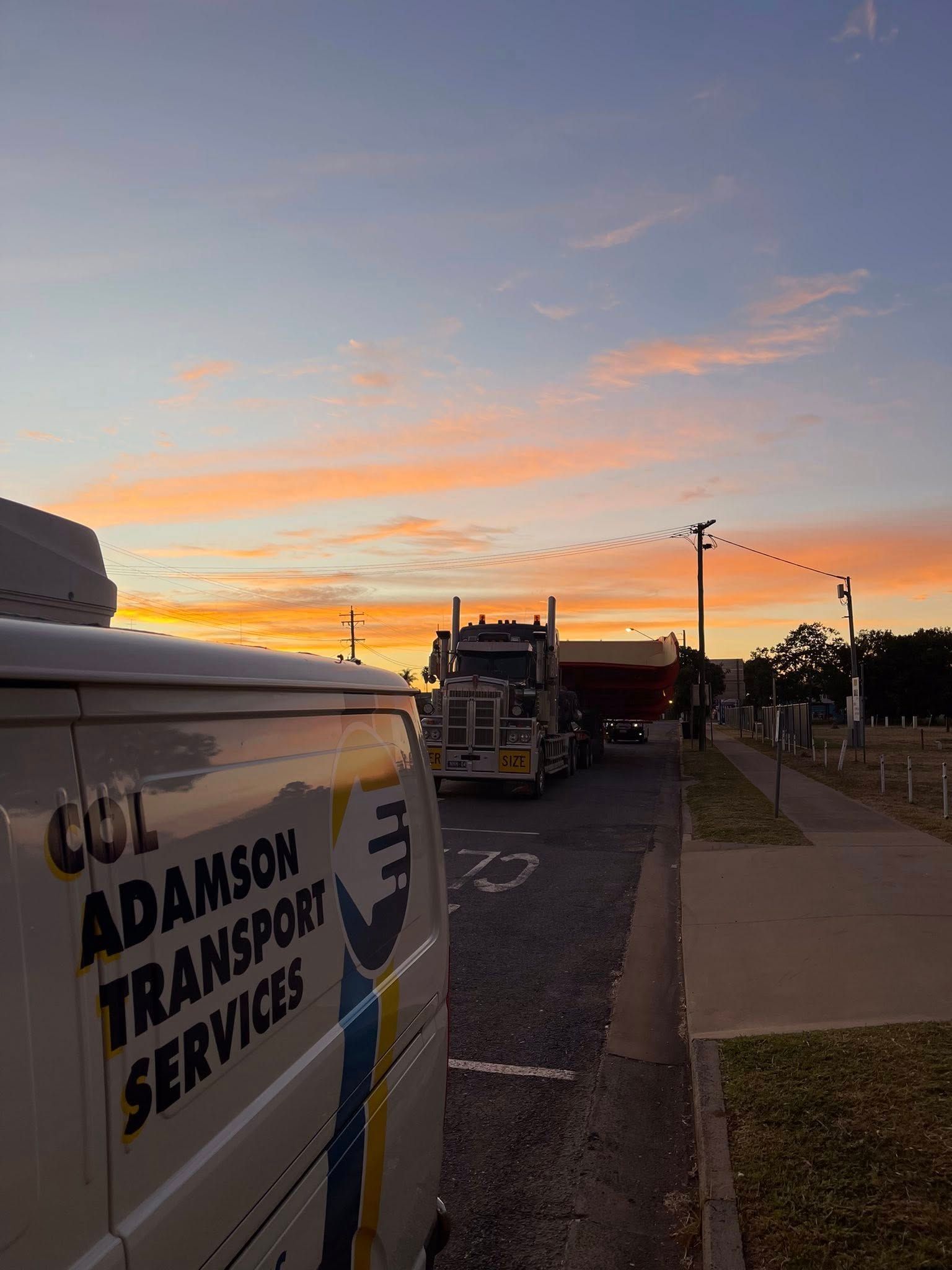 A large transport truck parked on a road at sunset, viewed beside a white van branded with Adamson Transport Services — Col Adamson Transport Services in Farleigh, QLD