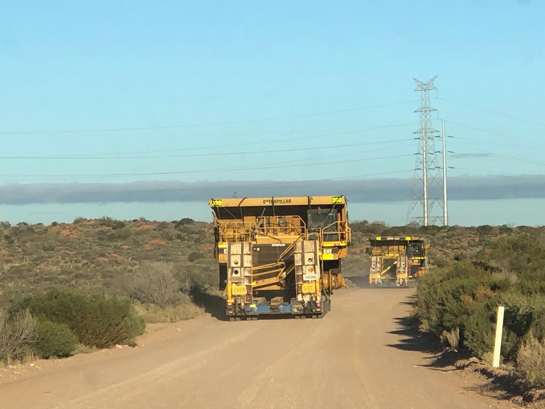 Two Large Yellow Mining Trucks Driving on a Dirt Road in a Desert Landscape — Col Adamson Transport Services in Farleigh, QLD