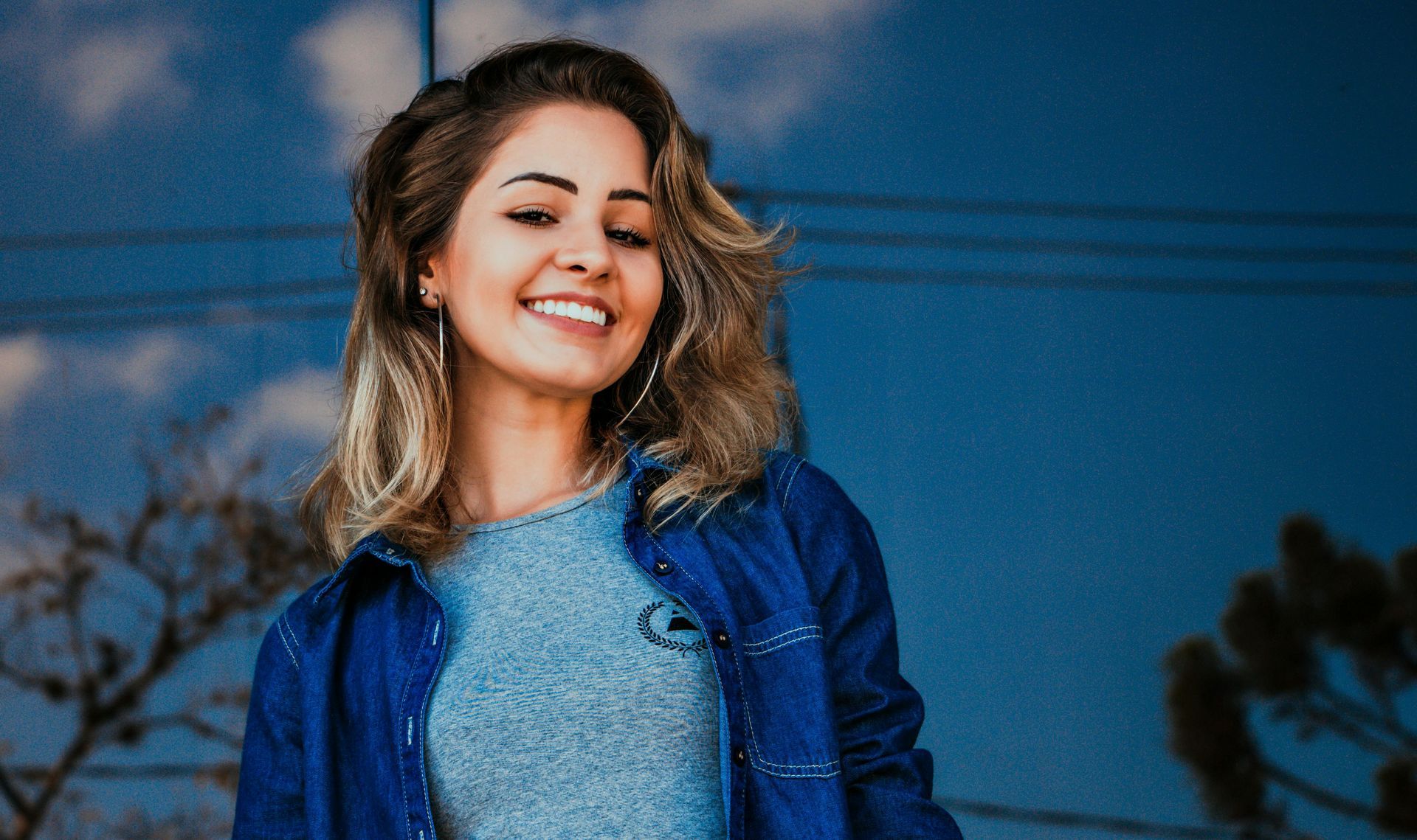Smiling woman in a blue jacket and gray shirt outdoors against a blue sky