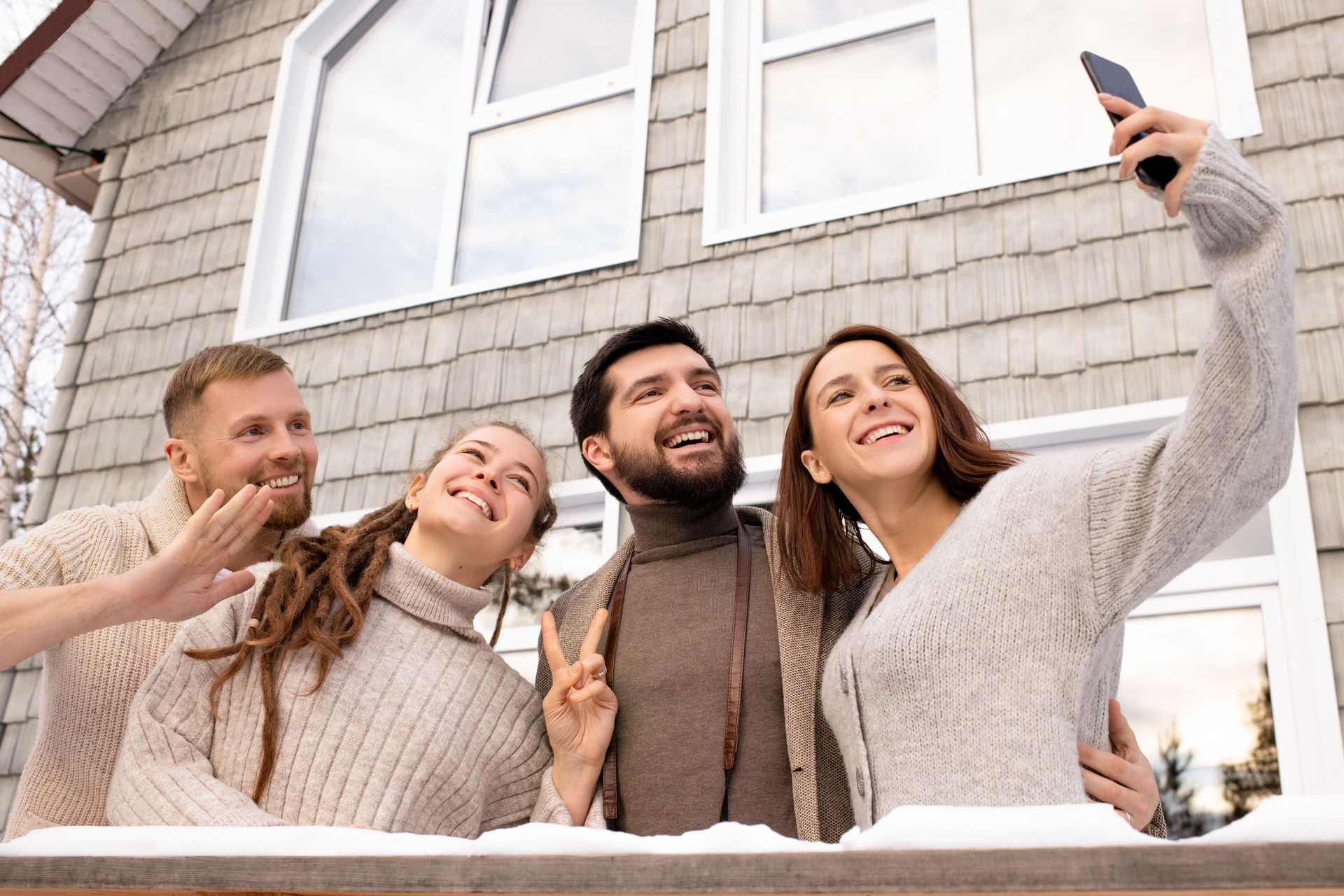 Four friends in winter sweaters smiling and taking a selfie outside a house, waving from behind a snowy railing