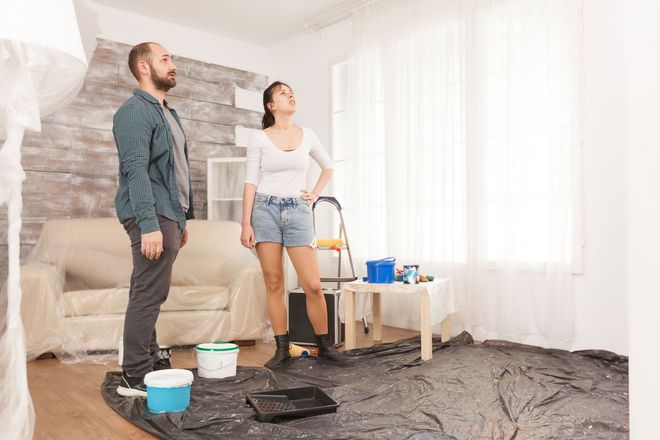 Two people standing in a room covered with drop cloths, painting supplies nearby and a ladder by the window
