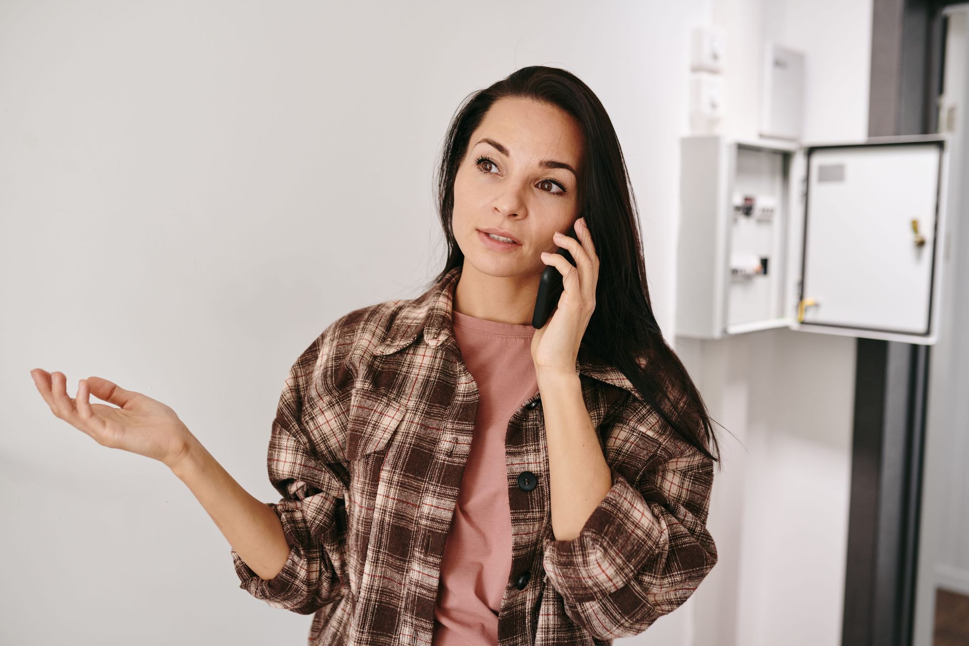 Woman on a phone call in a kitchen, wearing a pink shirt and plaid overshirt, gesturing with her free hand.
