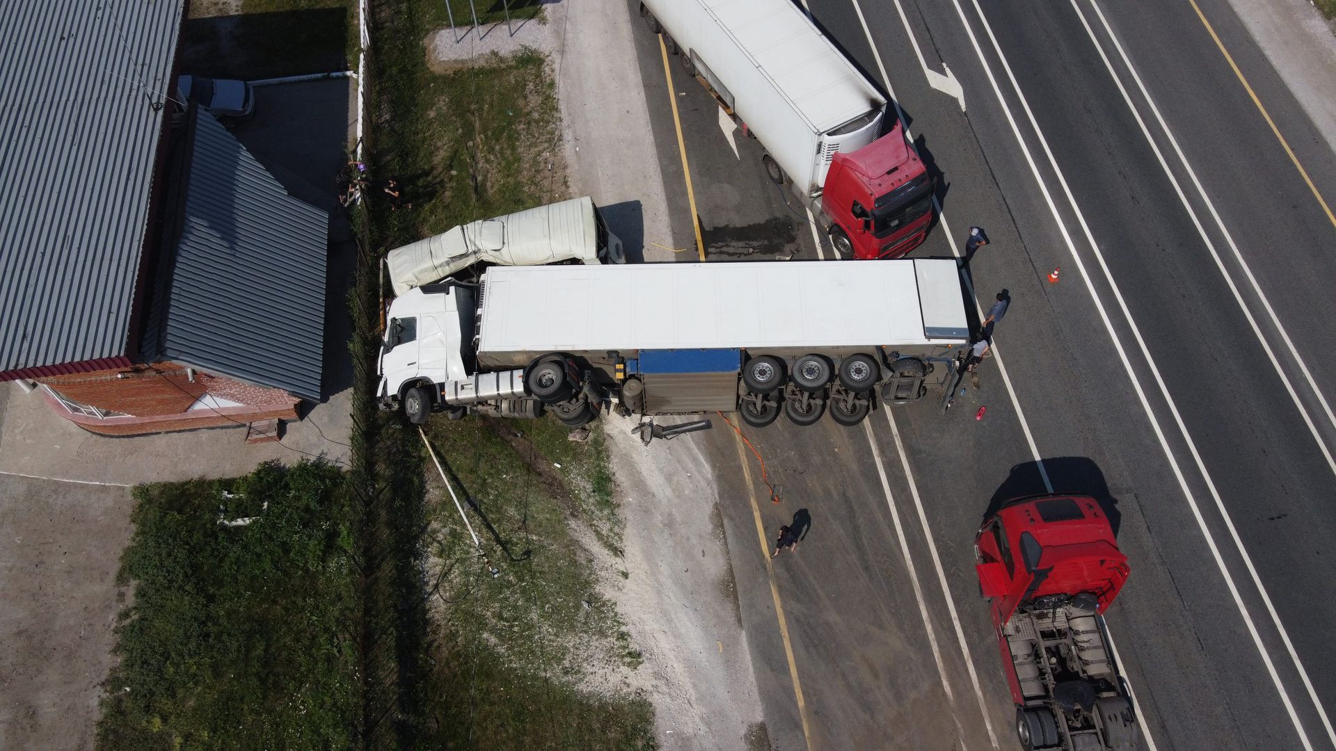 Aerial view of a white semi-trailer truck jackknifed on a road beside a red car and a building