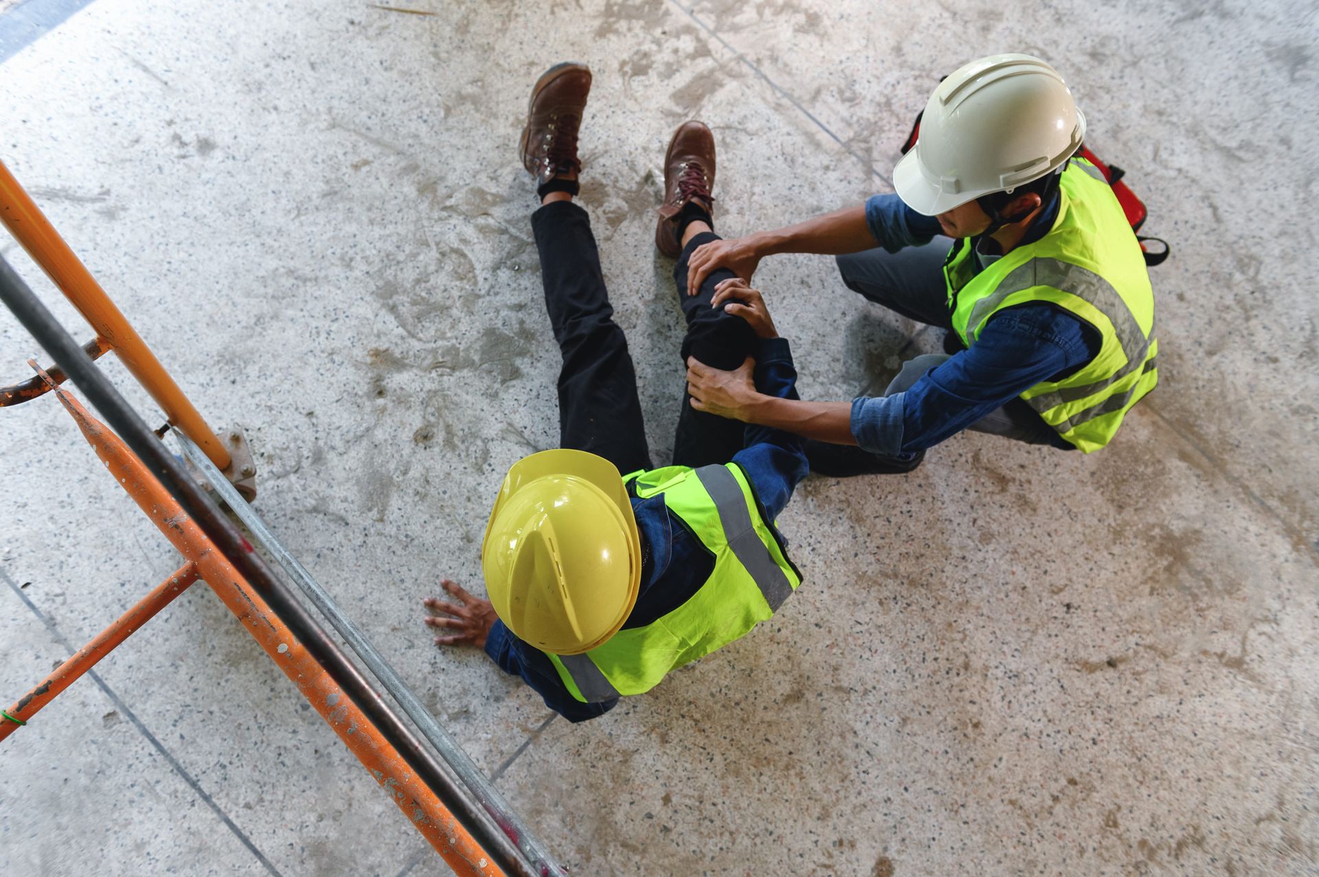 Two construction workers in hard hats and safety vests sit on a concrete floor, one checking the other's leg.