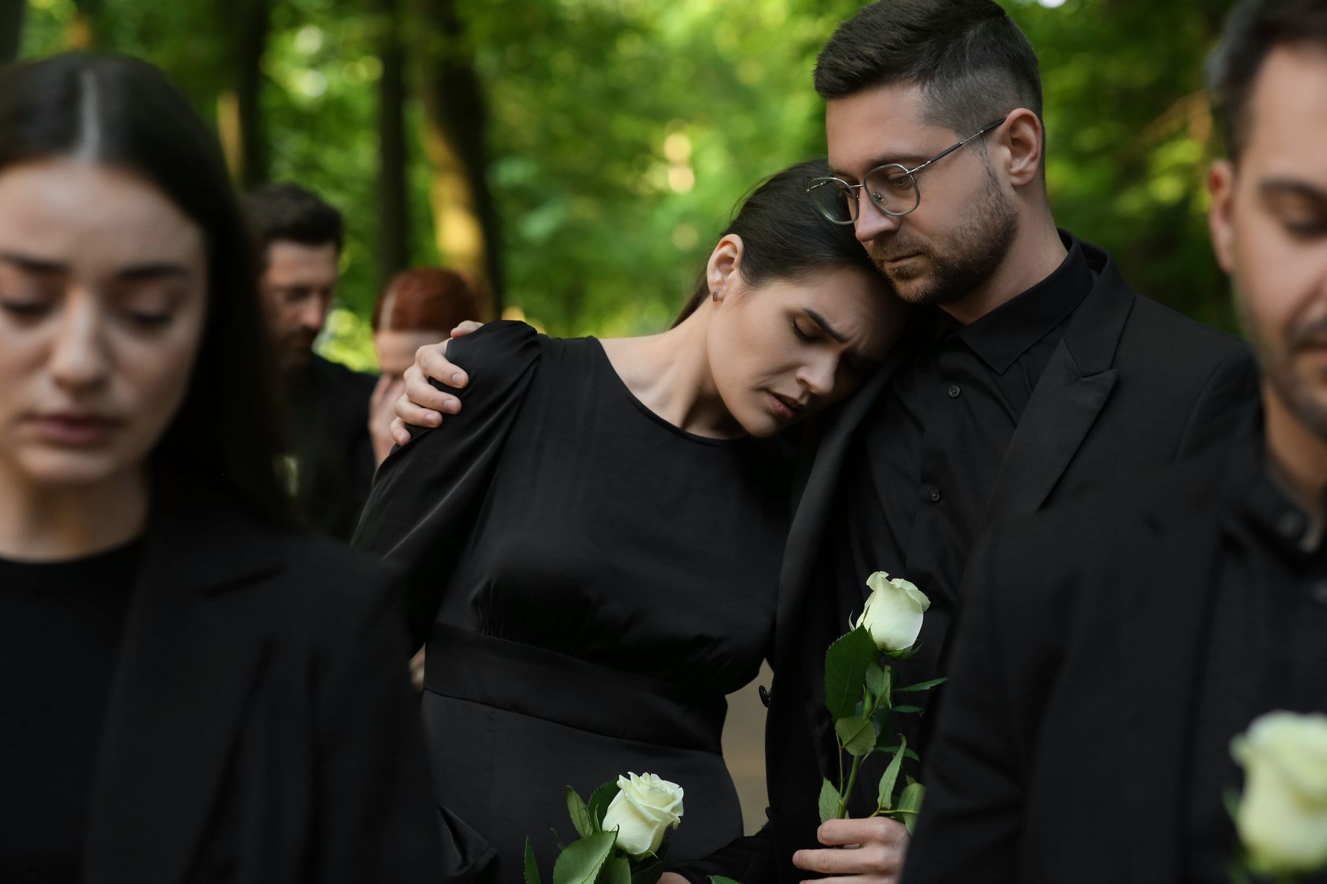 Mourners in black stand close together outdoors, one person resting their head on another’s shoulder, white flowers visible