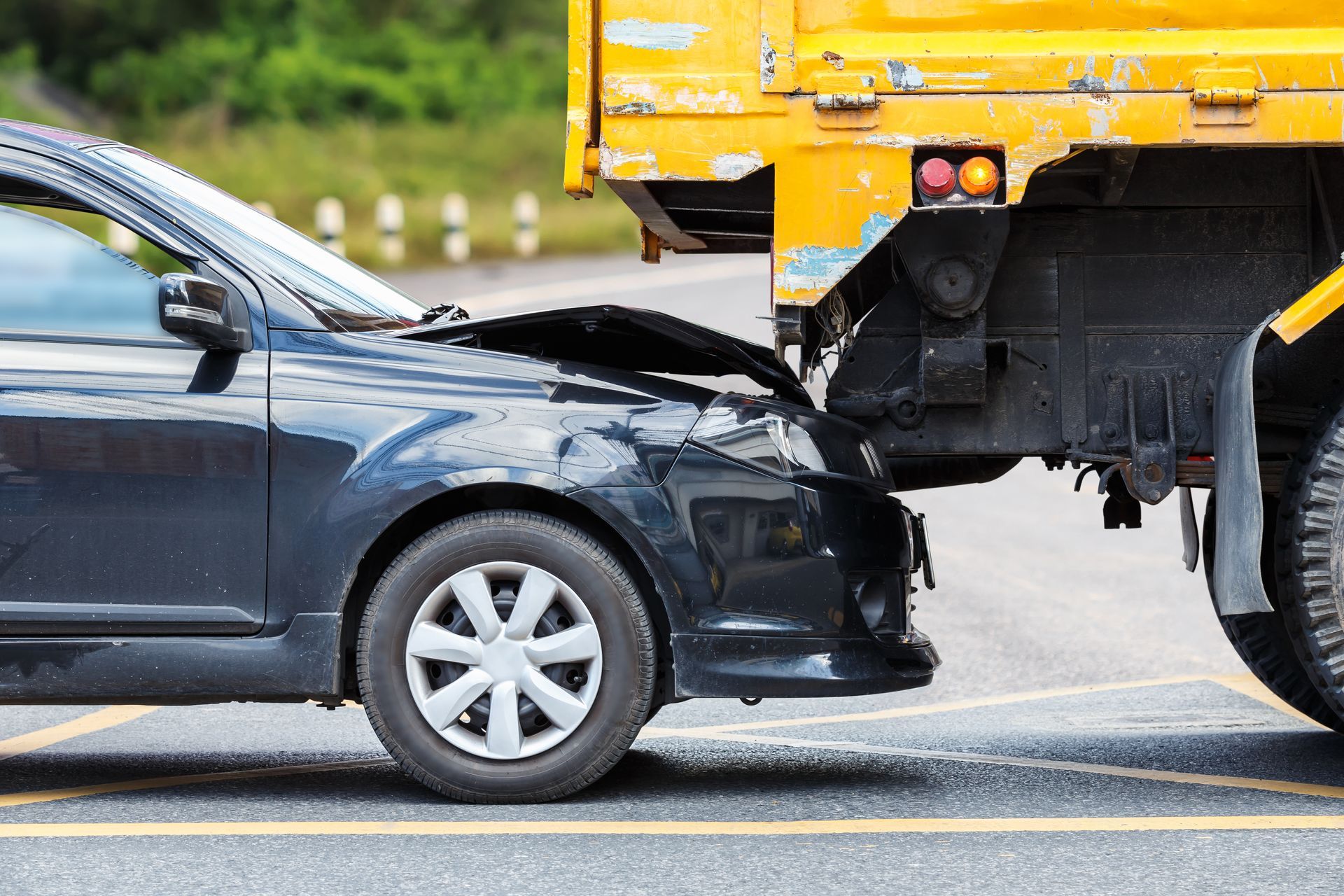 Black car crashed into the back of a yellow truck. The car's front is heavily damaged, on a paved road.