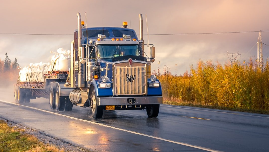 Semi-truck driving on a wet road, carrying cargo, under a cloudy sky.