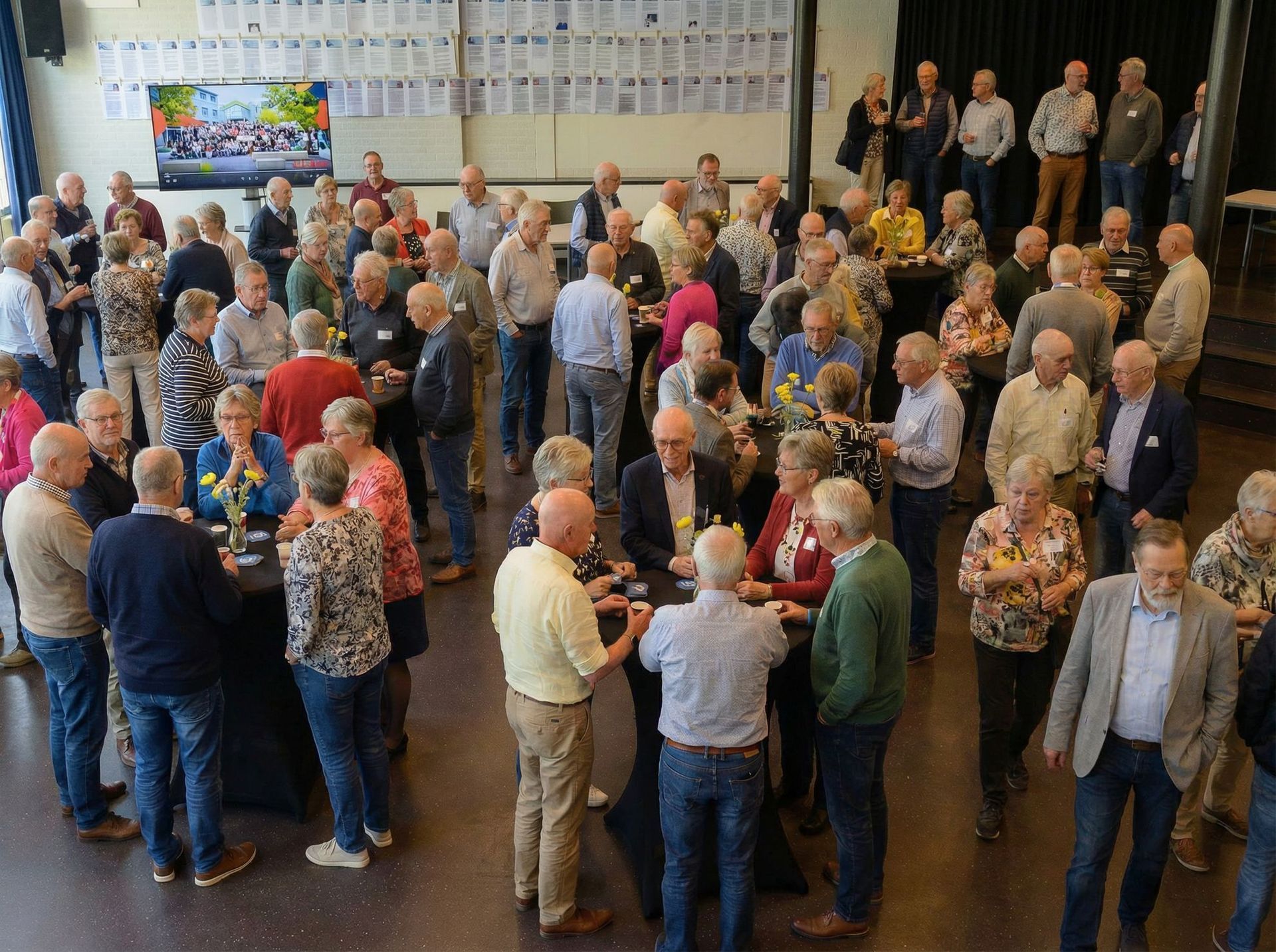 Een drukke receptie binnen met veel mensen die praten en in kleine groepjes bij een podium of presentatieruimte staan.