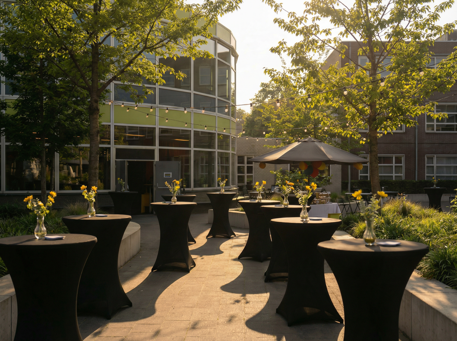 Buitenpatio met zwarte cocktailtafels en potplanten naast een glazen gebouw in het warme zonlicht.