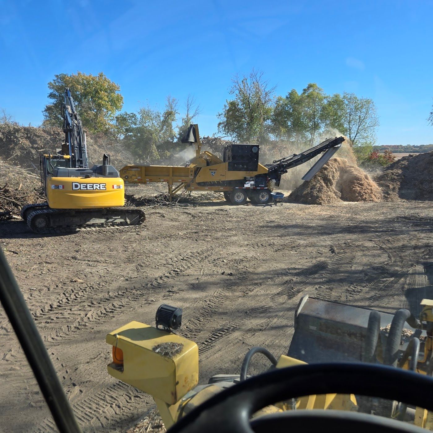 A John Deere excavator and wood grinder operating in a dusty, sunlit outdoor clearing.