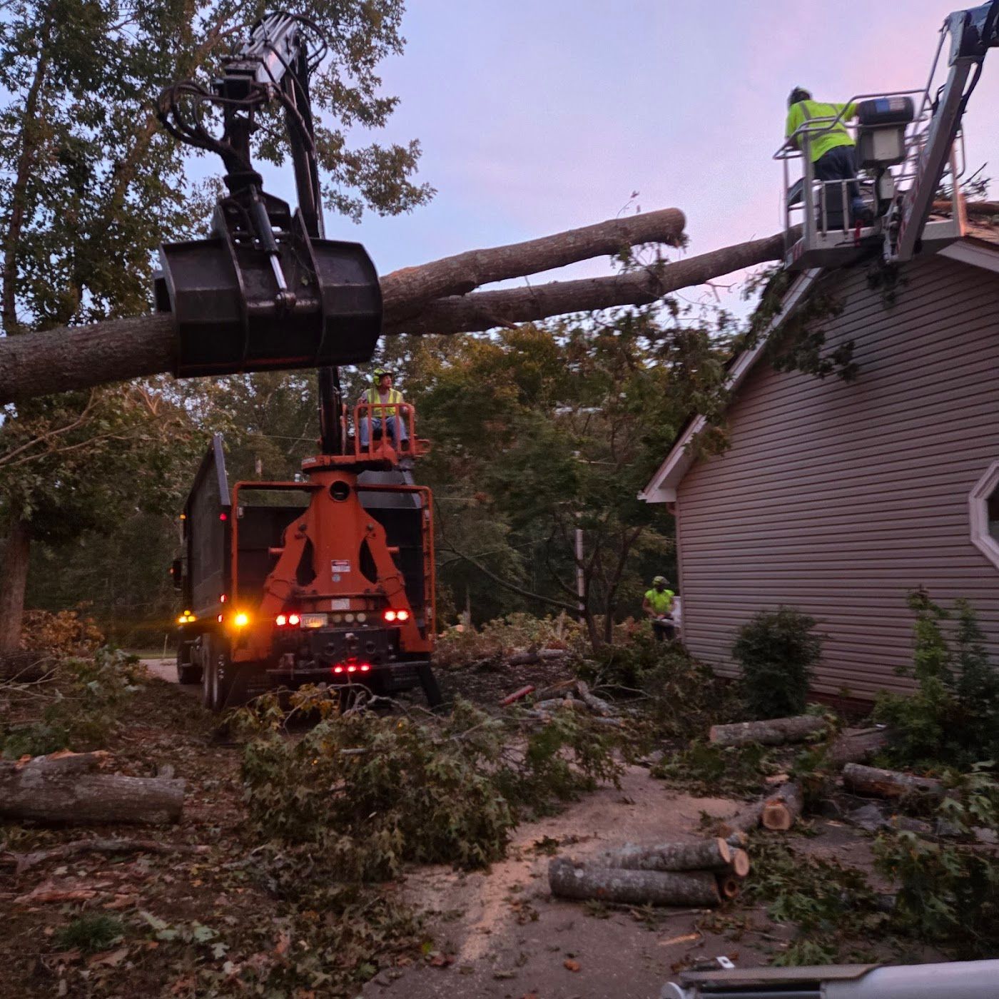 Tree removal crew using a knuckle boom loader to lift a large fallen tree limb away from a house roof at dusk.
