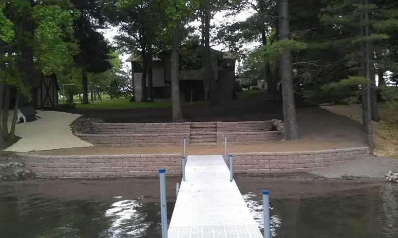 A lakefront property featuring newly installed tiered stone retaining walls leading to a white dock extending into the water.