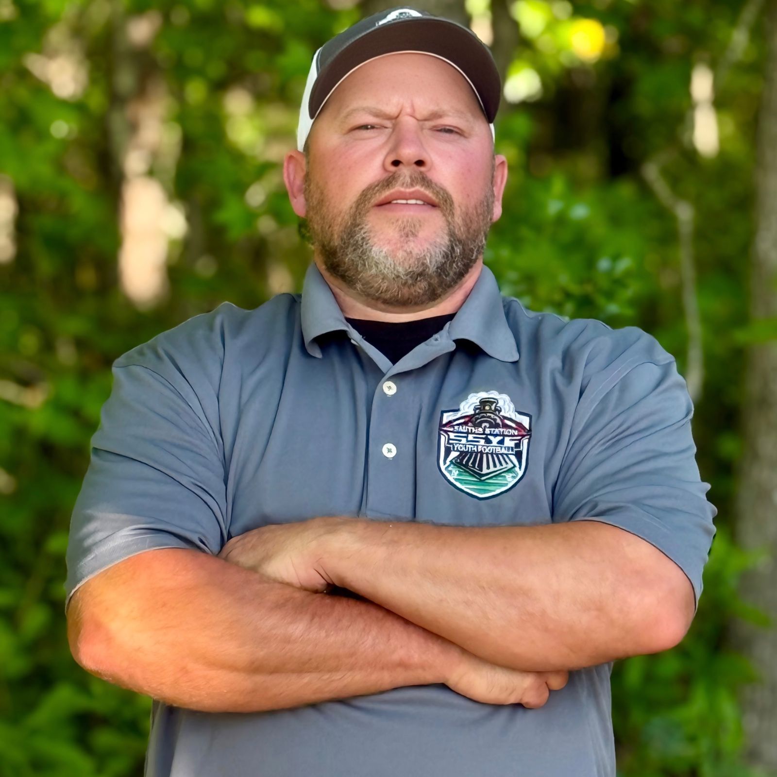 Man with crossed arms on a football field. He is wearing a blue polo shirt, khaki pants, and has a serious expression.