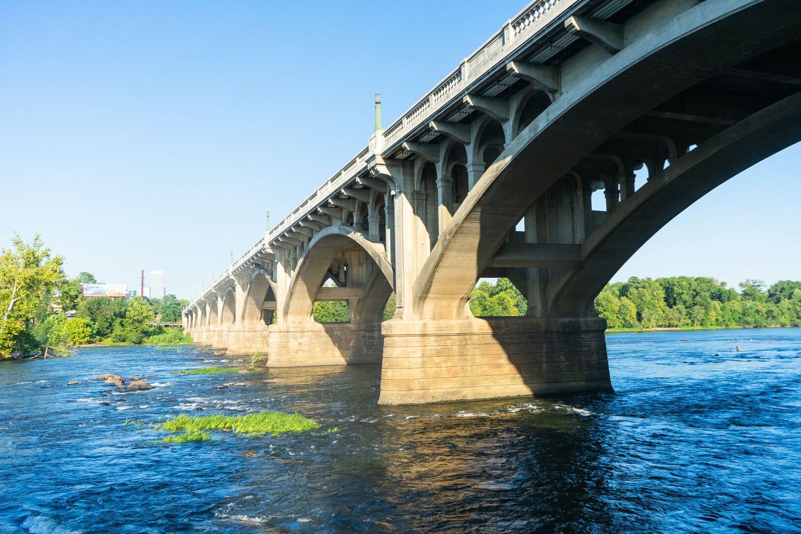 Bridge spanning a river; concrete arches support the road above. Green trees line the banks, blue water.