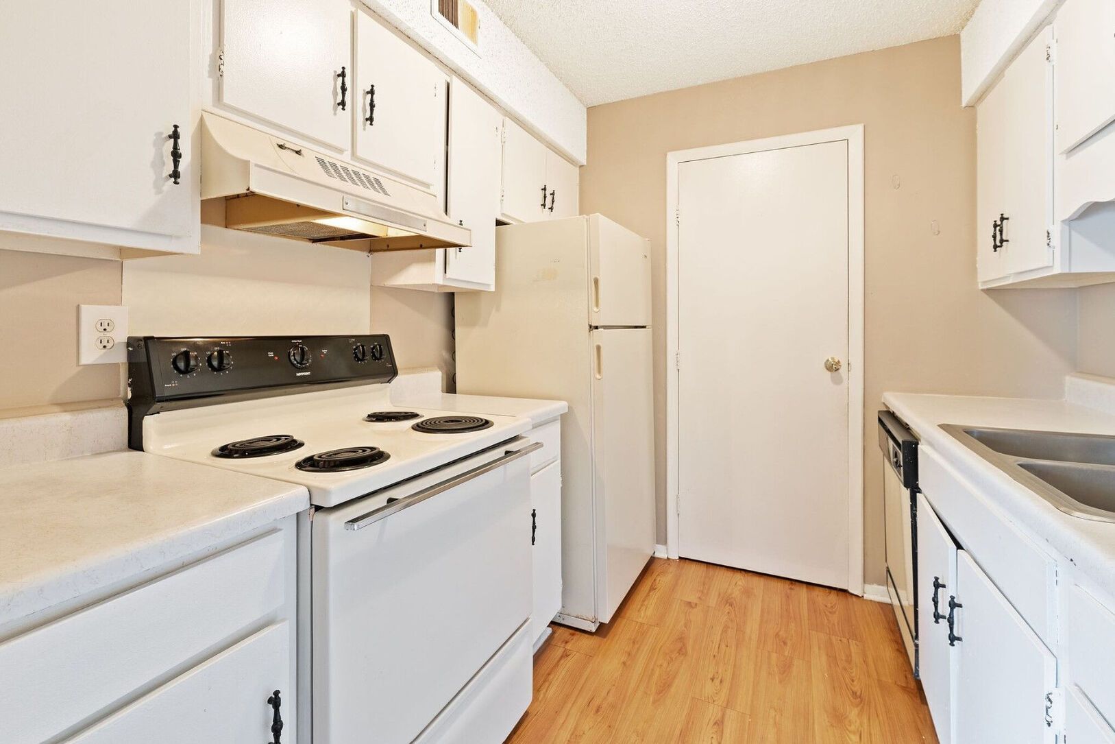 A small, white kitchen with a stove, refrigerator, cabinets, and a door. Wooden floors and beige walls are visible.