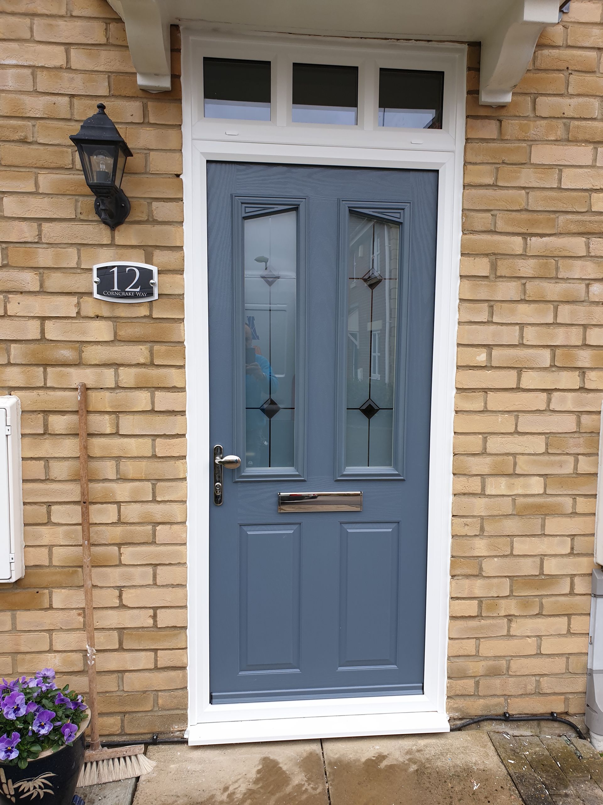 Blue front door on a brick house, with a black lantern, house number 12, and potted flowers nearby.