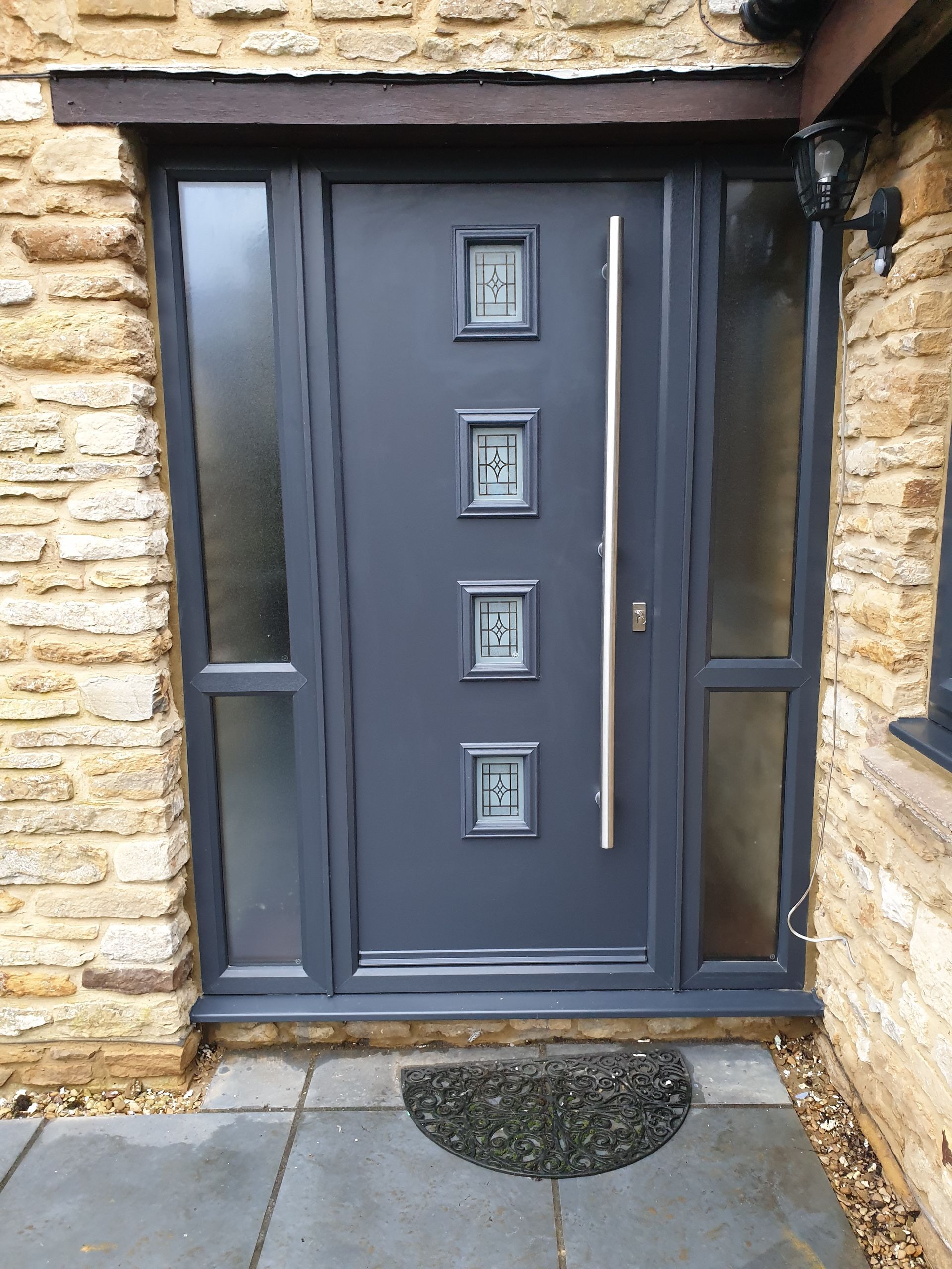 Dark gray front door with four small windows, set in a stone wall beside a narrow sidelight and doormat.