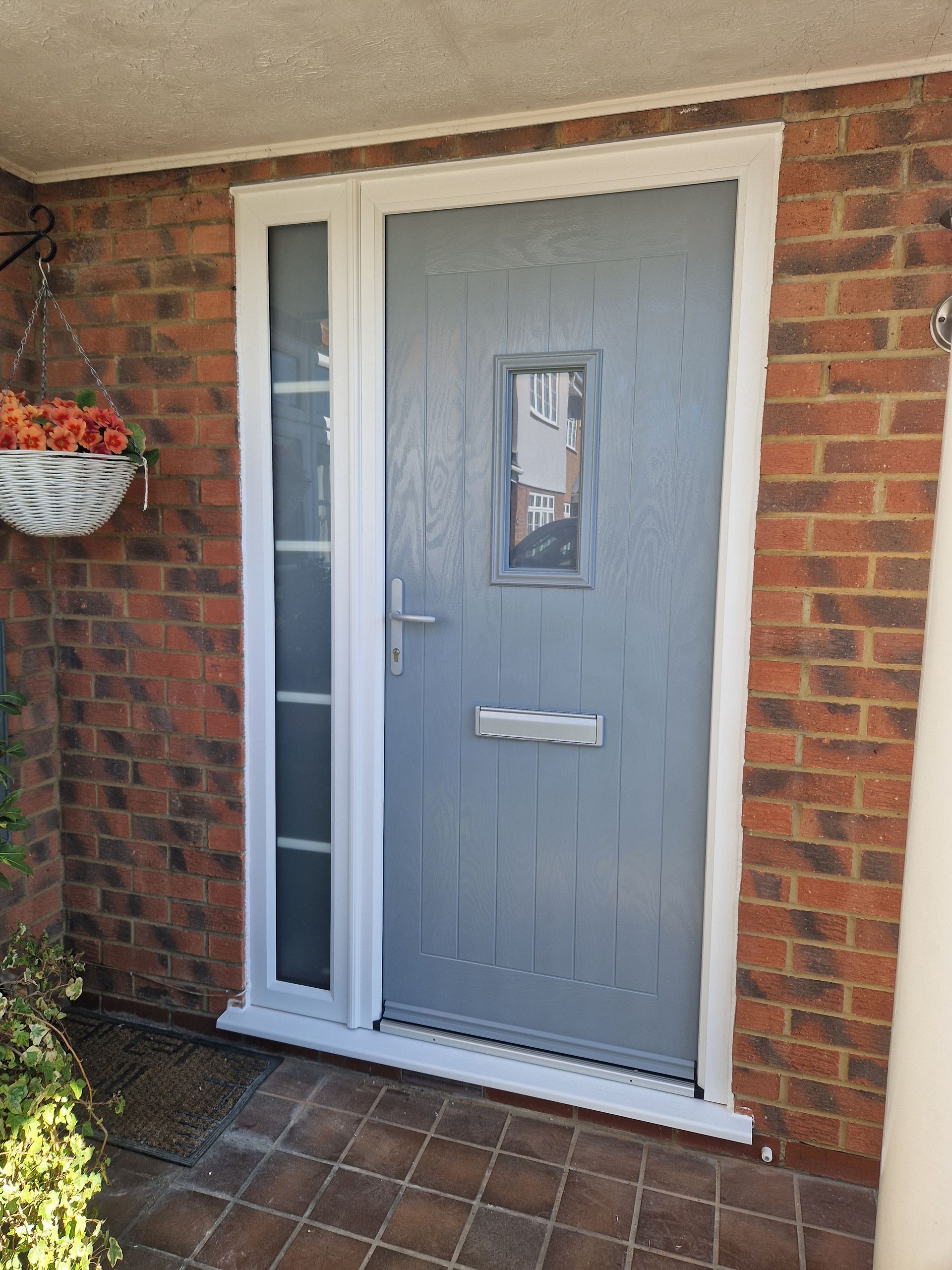 Gray front door with narrow sidelights set in a red brick wall, beside a hanging flower basket.