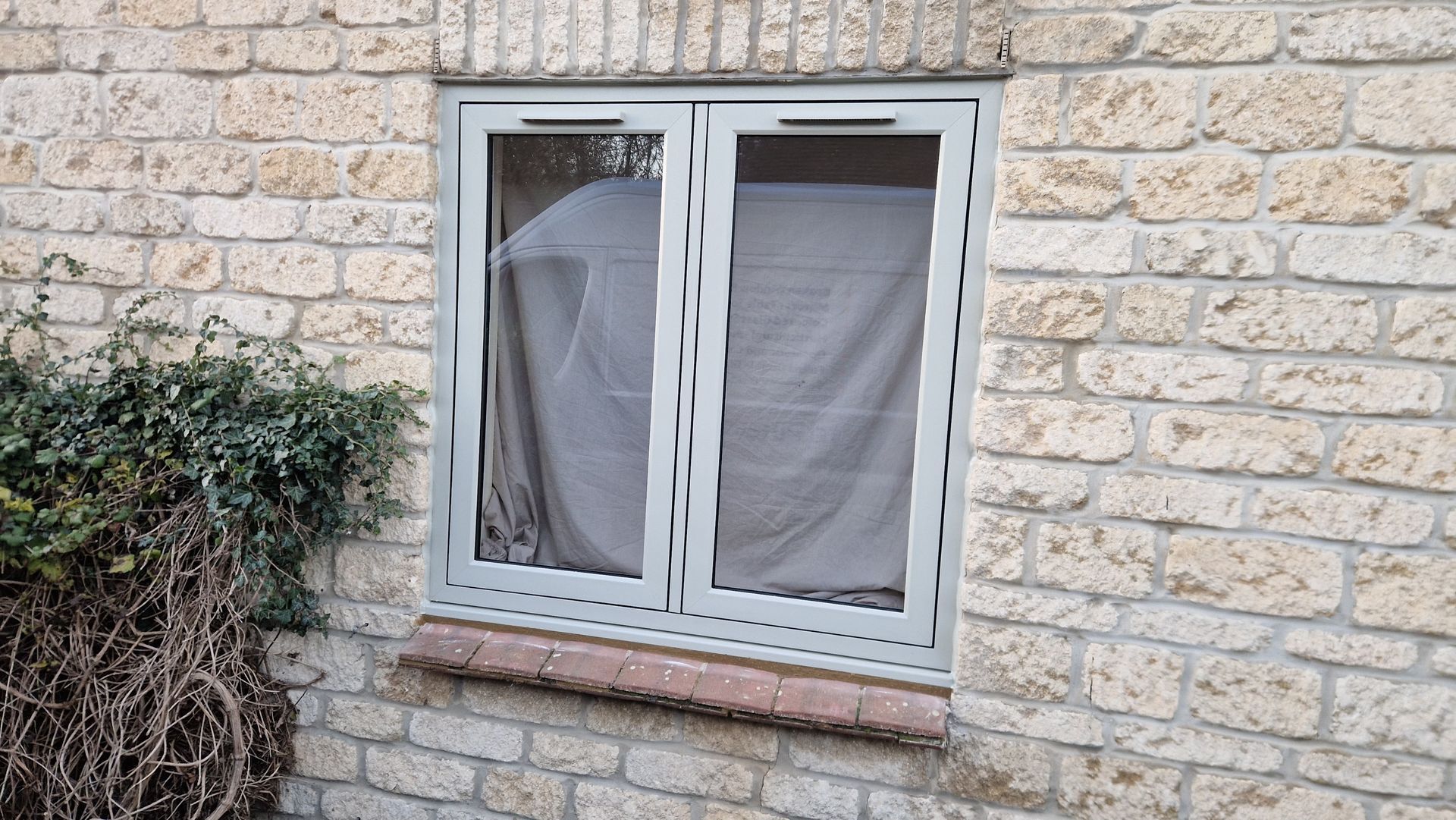 Gray-framed window set in a stone wall, covered with white curtains behind the glass.