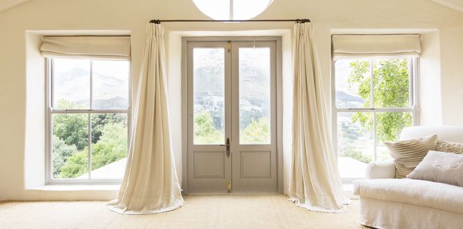 Bright beige living room with French doors, sheer curtains, and greenery visible through large windows.