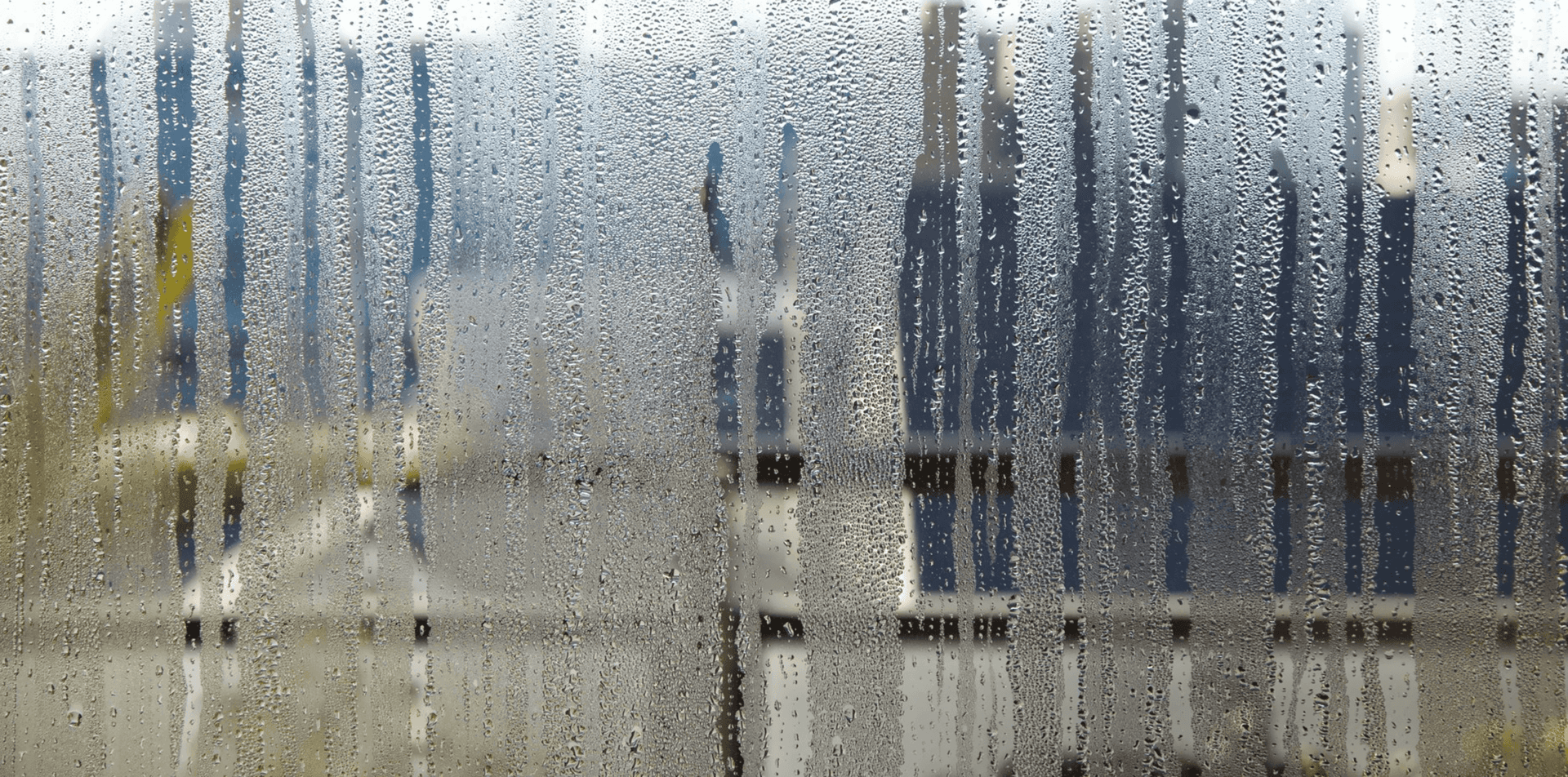 Blurred view of a window with raindrops and vertical blinds, showing buildings outside.