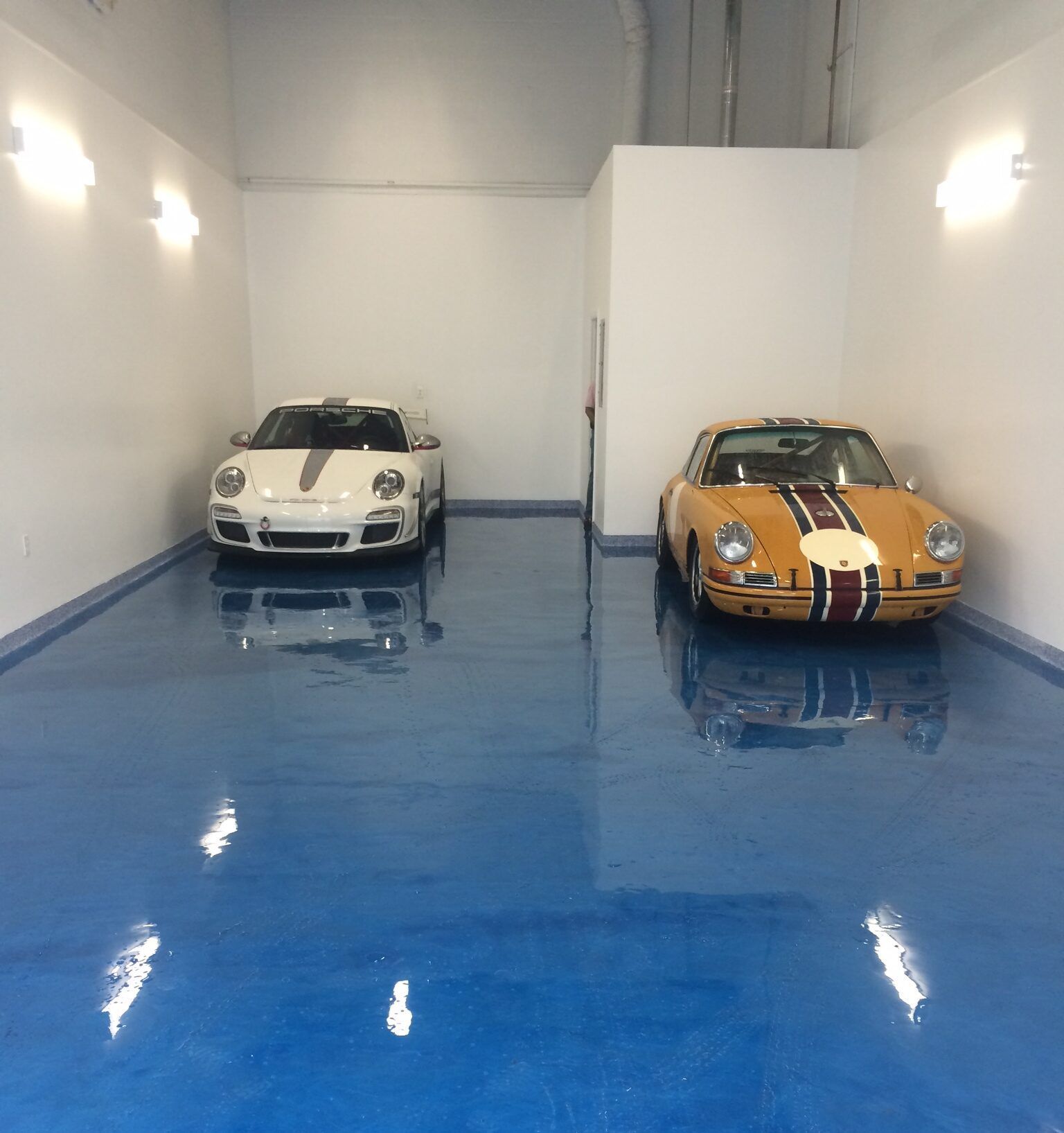 Two vintage and modern Porsche sports cars parked in a brightly lit garage with a polished, reflective blue epoxy floor.