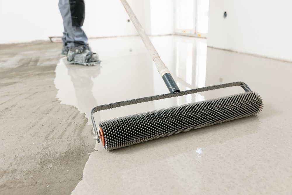 A worker uses a spiked roller to smooth wet self-leveling compound on a concrete floor.