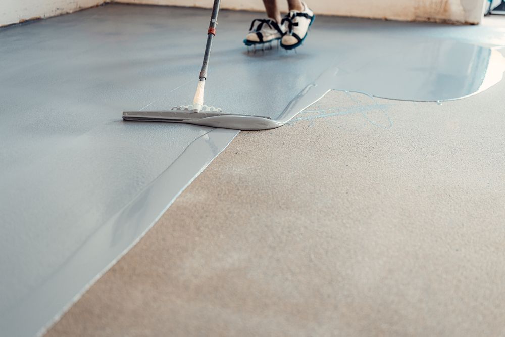 A person wearing spiked shoes uses a squeegee to spread smooth gray epoxy coating over a concrete floor.