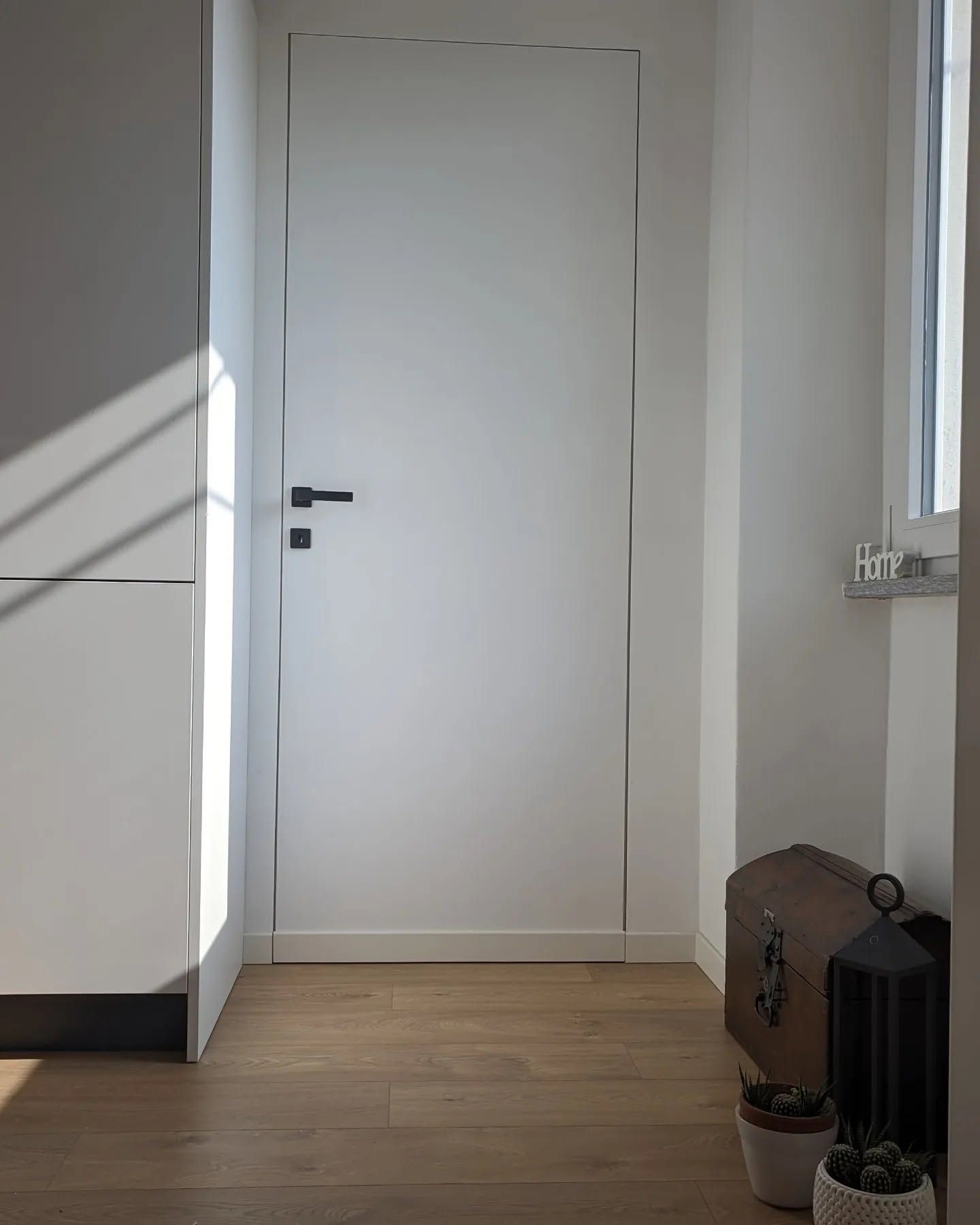 Ingresso minimalista con una porta bianca, luce proveniente dalla finestra e un piccolo mobile marrone accanto a una lanterna