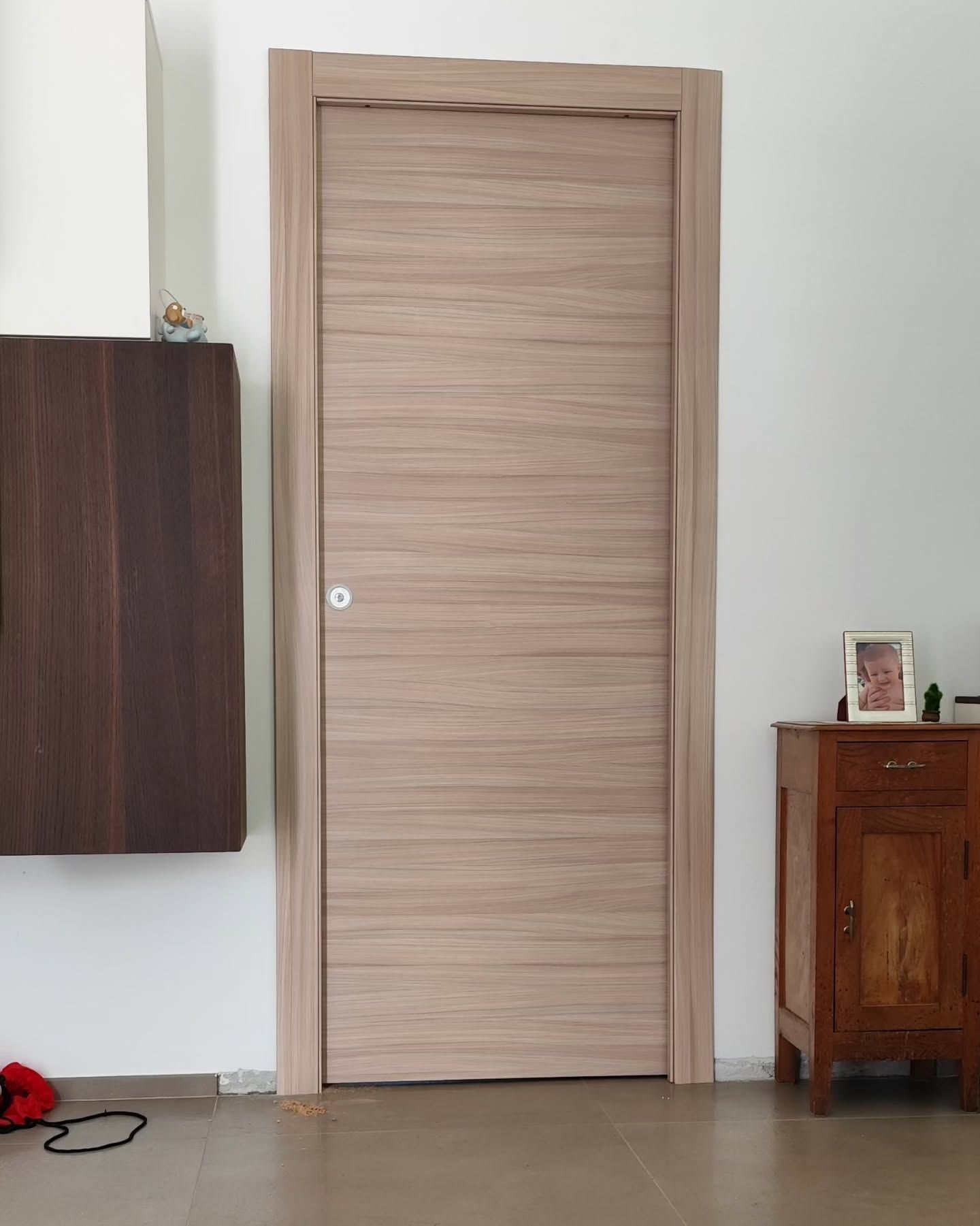 Porta interna in legno chiaro al centro di una stanza piccola, affiancata da un mobile scuro e da un tavolino in legno.