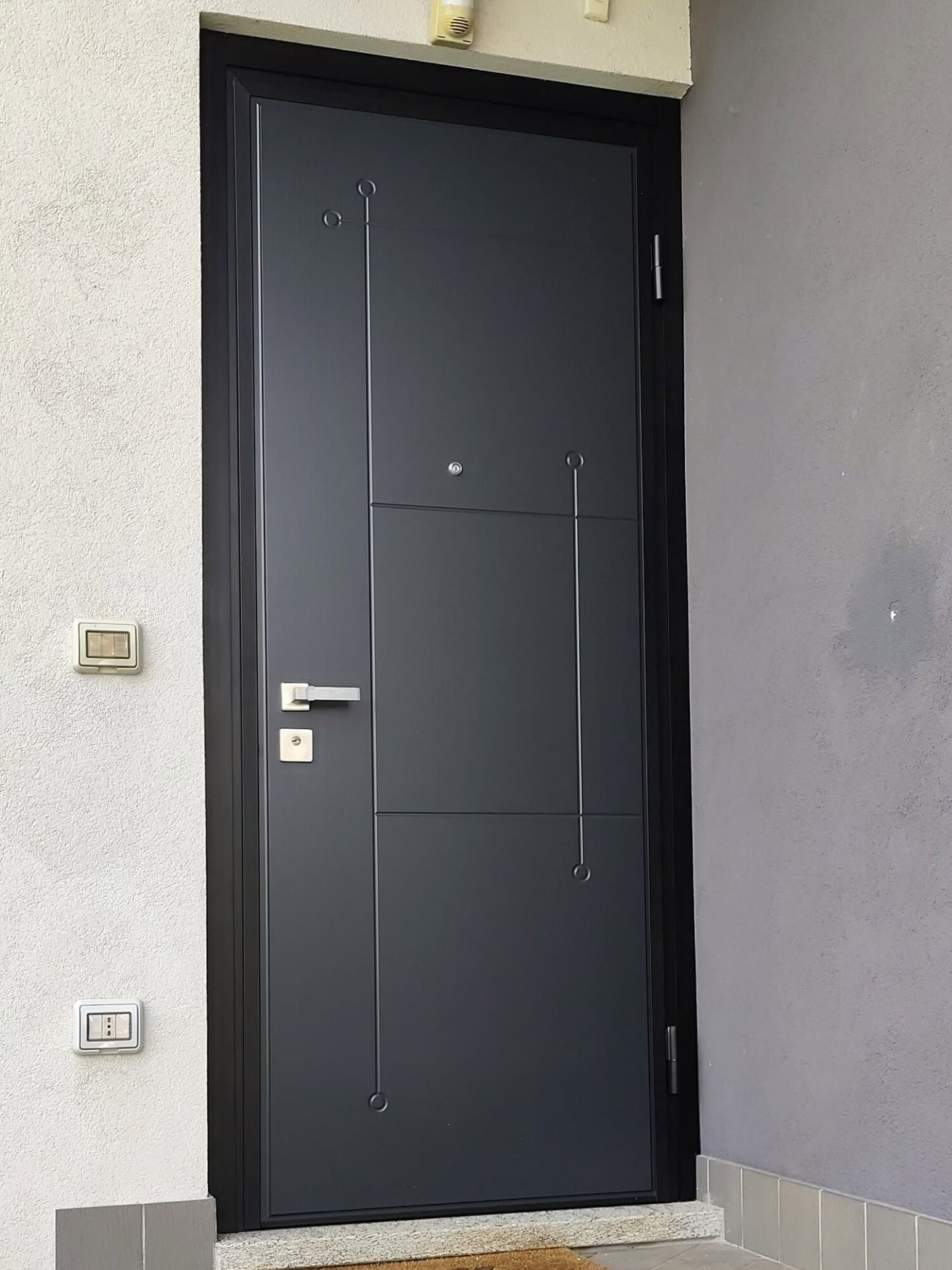 Porta d’ingresso moderna nera con maniglia argentata, inserita in una parete esterna di colore chiaro.