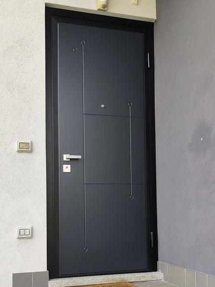 Porta d’ingresso moderna nera con maniglia argentata, inserita in una parete esterna di colore chiaro.