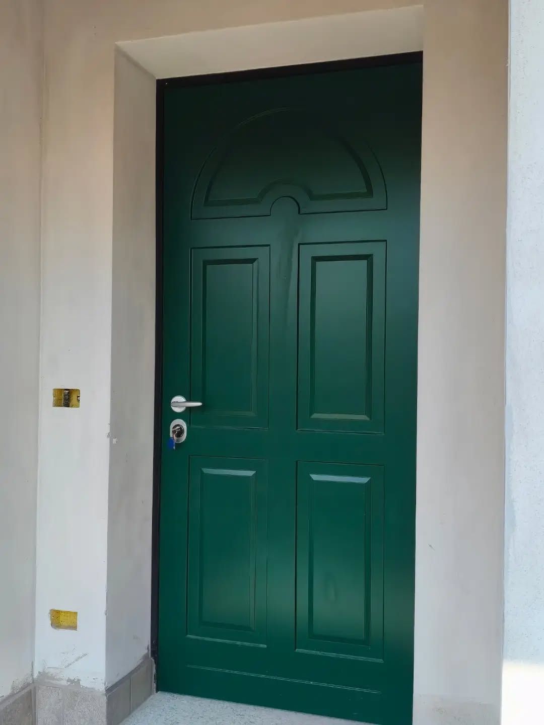 Porta d’ingresso verde con pannelli decorativi, incastonata in una parete chiara.